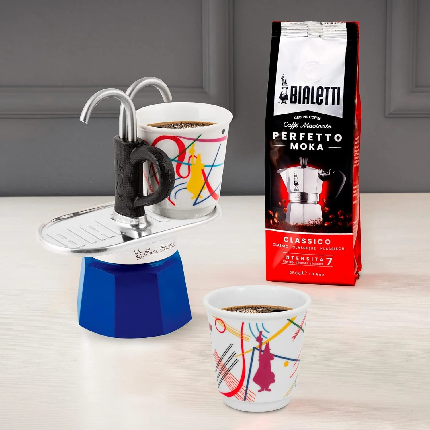 Una macchina per espresso blu e argento, due tazze di caffè e un sacchetto di chicchi di caffè sono esposti su una superficie bianca.