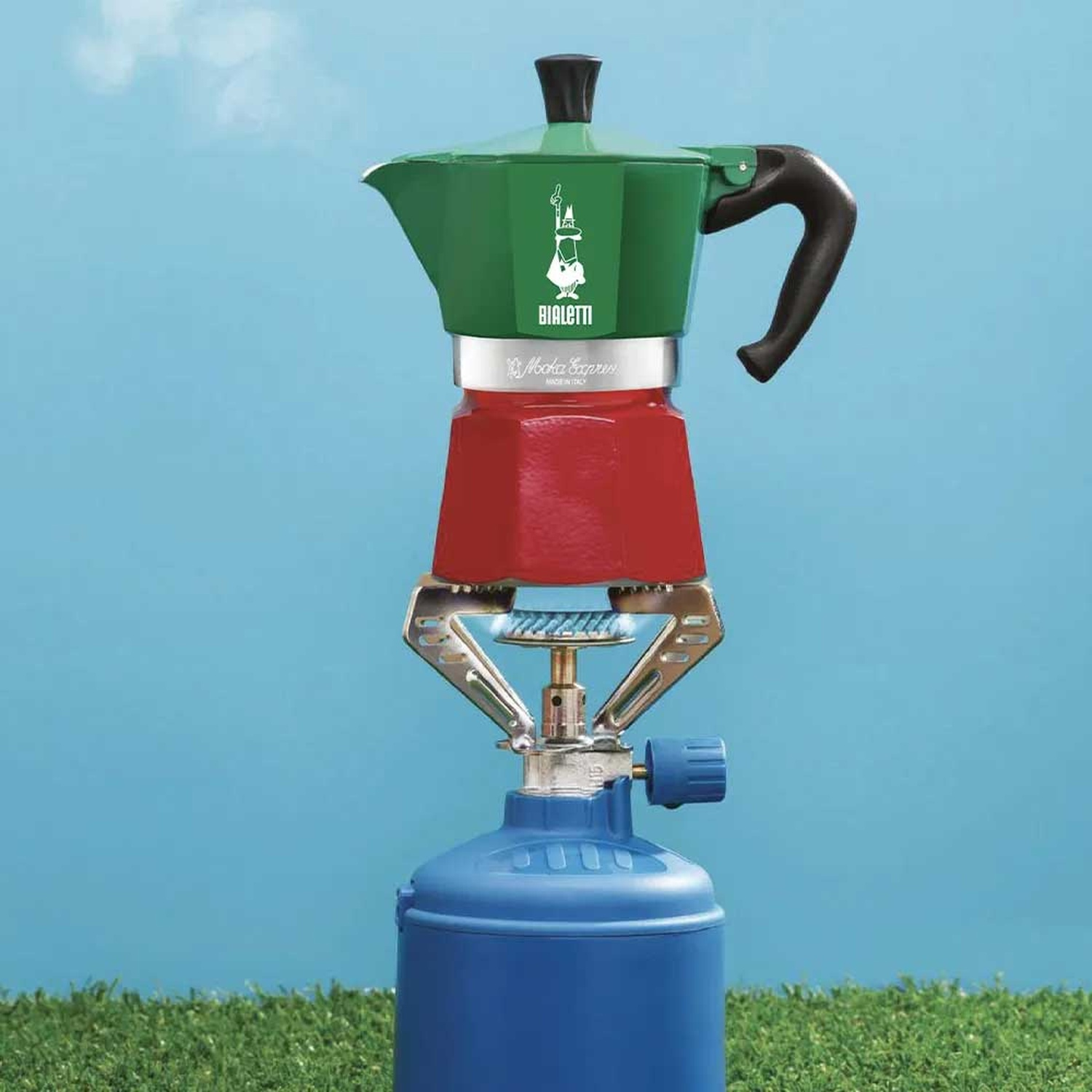 Una caffettiera Bialetti rossa e verde è posta su un fornello a gas blu, che produce vapore. Lo sfondo è cielo blu ed erba verde.
