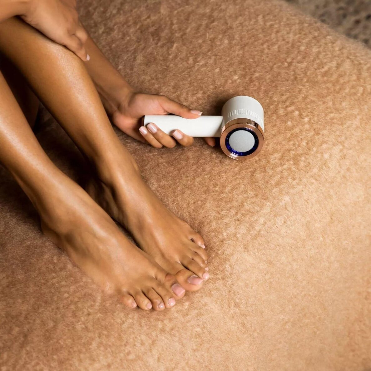 Osoba trzyma białą i złotą suszarkę do włosów w pobliżu nogi. Widoczne są stopy. Tło to brązowa, teksturowana powierzchnia.