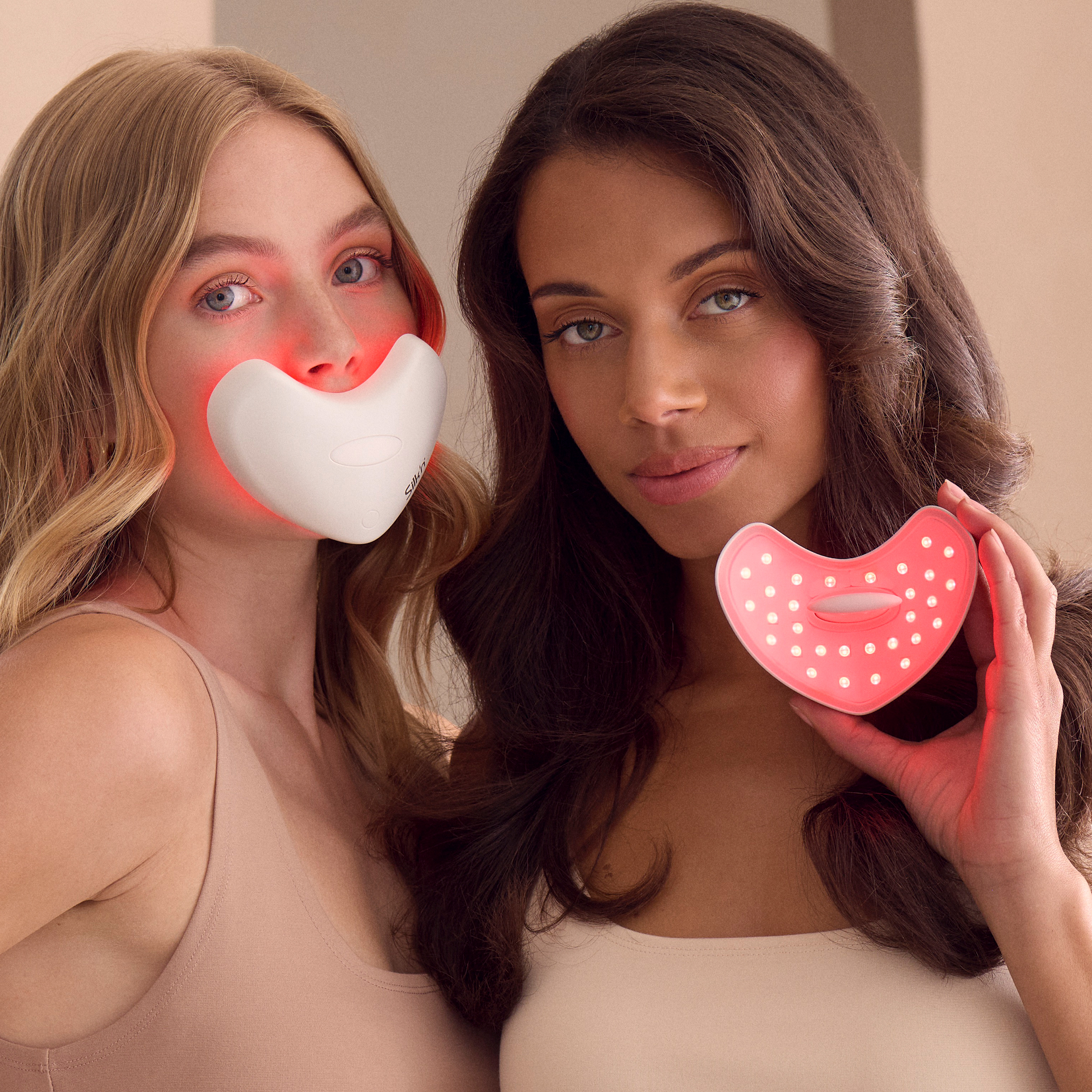 Dwie kobiety pozują z urządzeniami do terapii światłem czerwonym. Jedna ma na sobie maskę na twarz, druga trzyma urządzenie w kształcie serca.