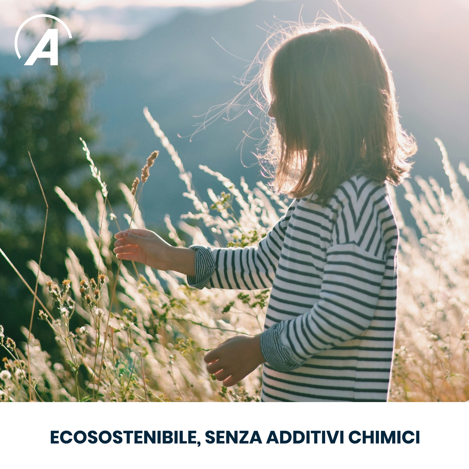 Un bambino con una maglietta a righe tocca erba alta. Le montagne sono sullo sfondo. Il sole splende e la scena è naturale.