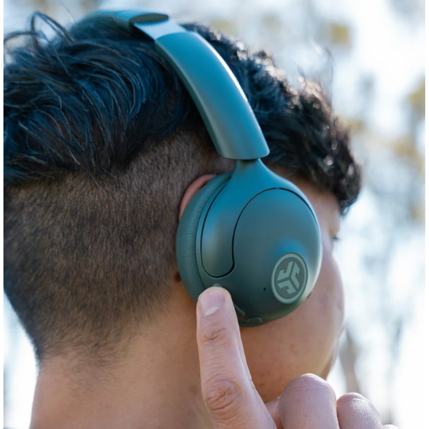 A person is wearing green headphones, and a finger is pressing a button. The background is blurred with light.