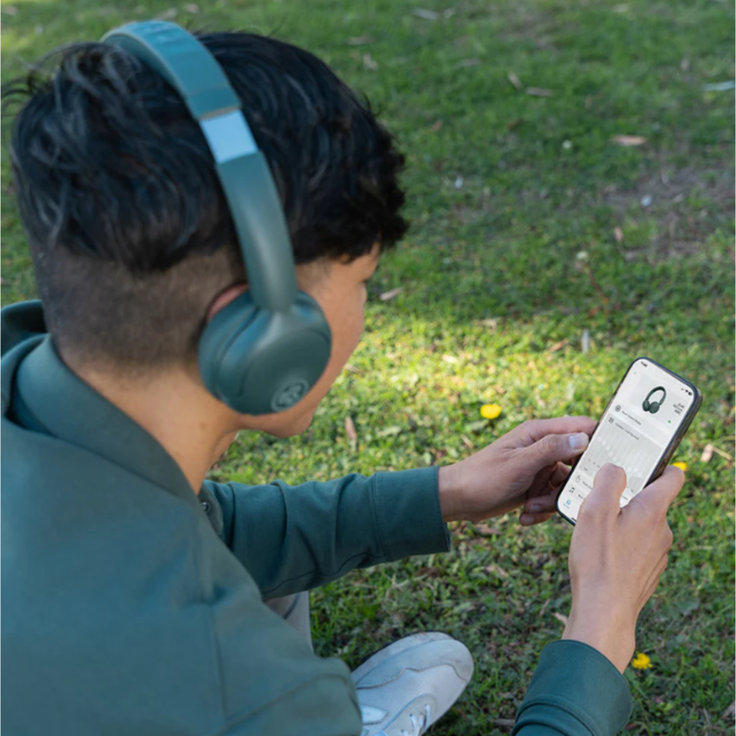 Person wearing headphones and looking at a phone outdoors. The phone displays an app interface. The background is grass.
