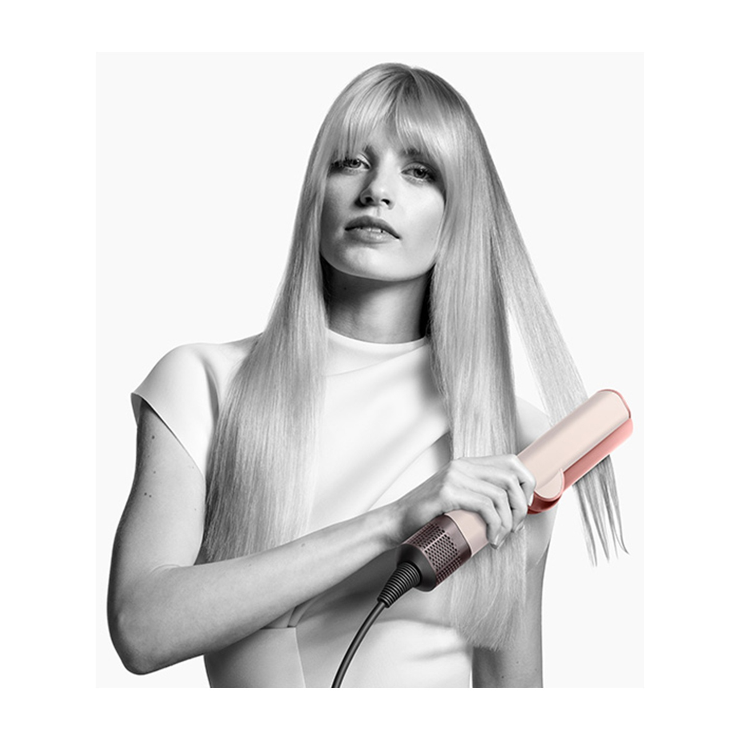 Donna con lunghi capelli biondi tiene in mano una piastra. Indossa un top bianco. Lo sfondo è bianco.