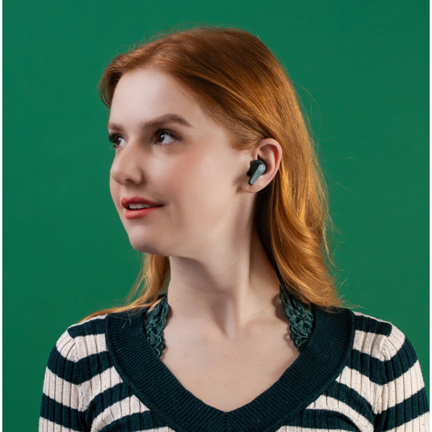 Une femme aux cheveux roux et un écouteur sourit, portant un pull rayé. Le fond est vert uni.