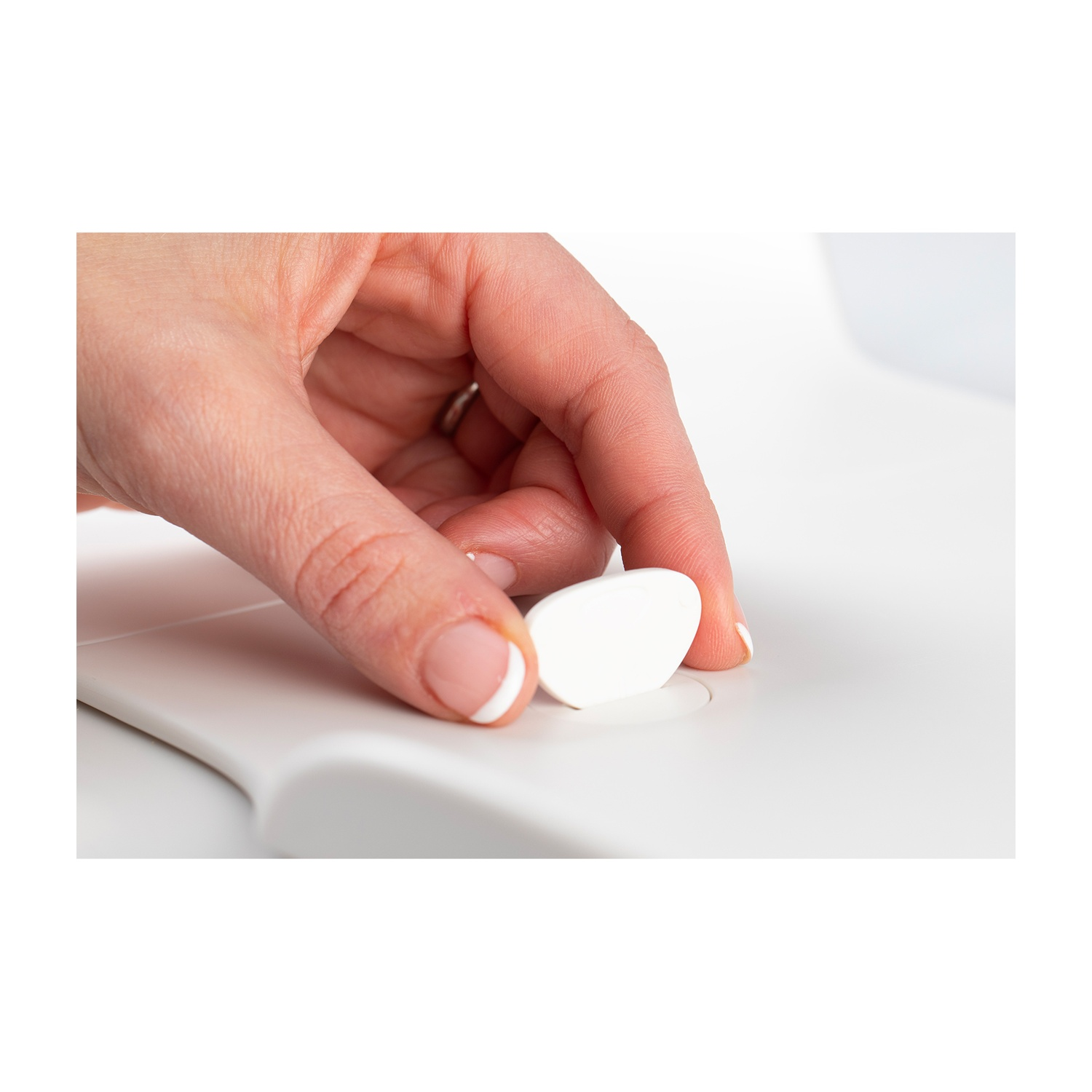 Una mano con una manicure alla francese preme un oggetto bianco ovale su una superficie bianca abbinata. C'è un anello al dito.