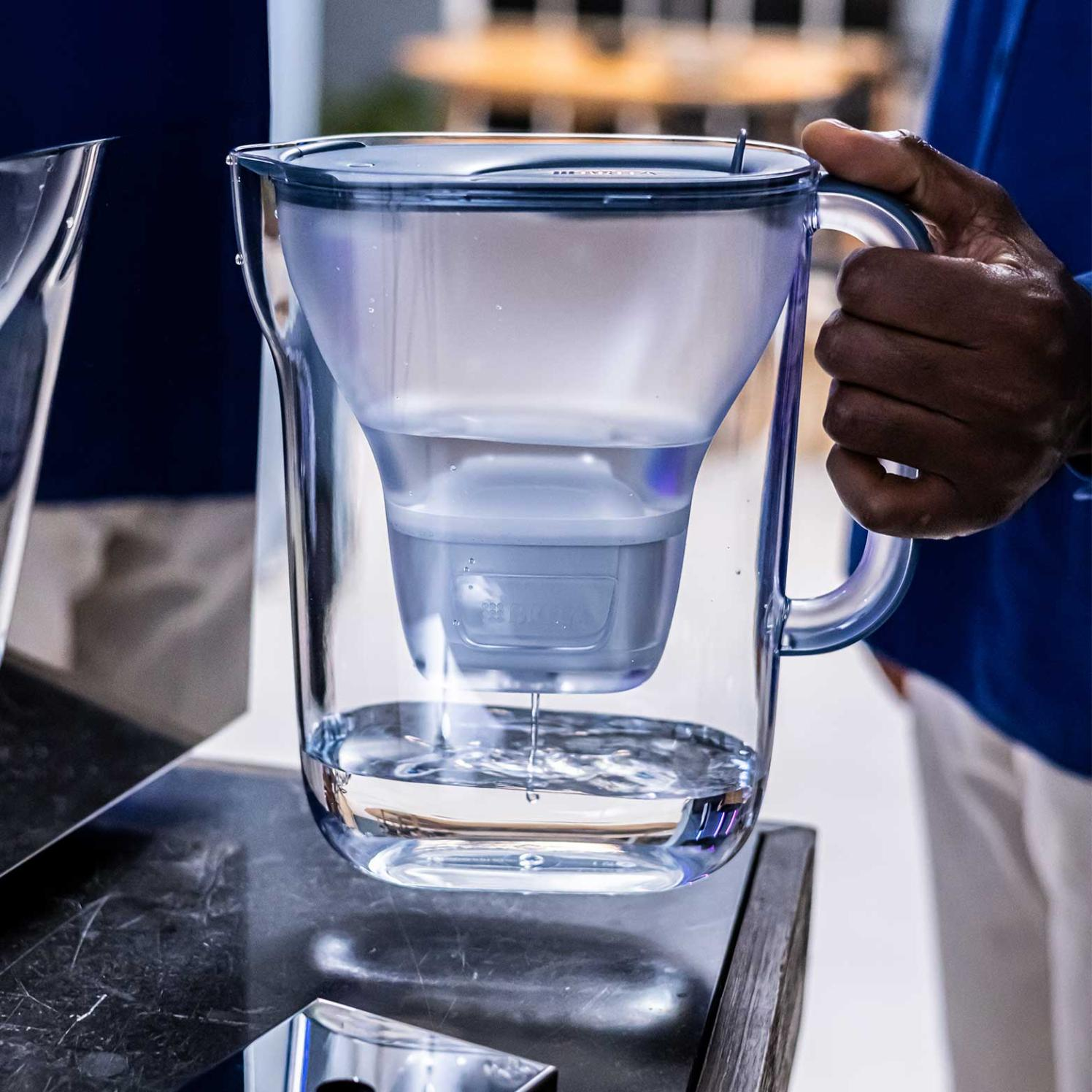 Ein klarer Wasserfilterkrug mit Wasser darin. Eine Hand hält den Griff. Wasser tropft aus dem Filter in den Krug.
