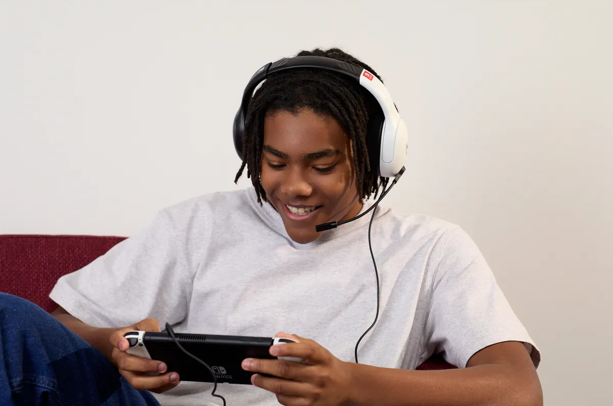 Ein lächelnder junger Mann mit Kopfhörern spielt mit einer Handheld-Konsole. Er trägt ein hellgraues Hemd und sitzt auf einem roten Sofa.