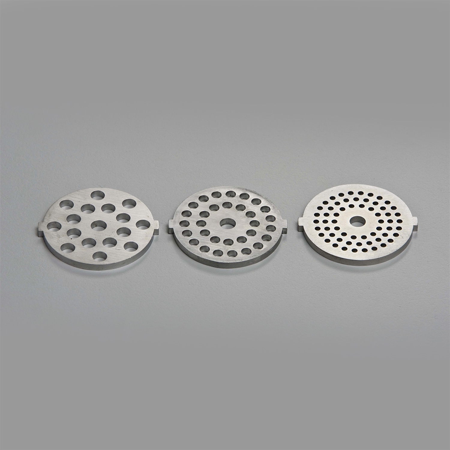 Tre piastre metalliche circolari con fori. Ogni piastra ha dimensioni e motivo dei fori diversi, su sfondo grigio.
