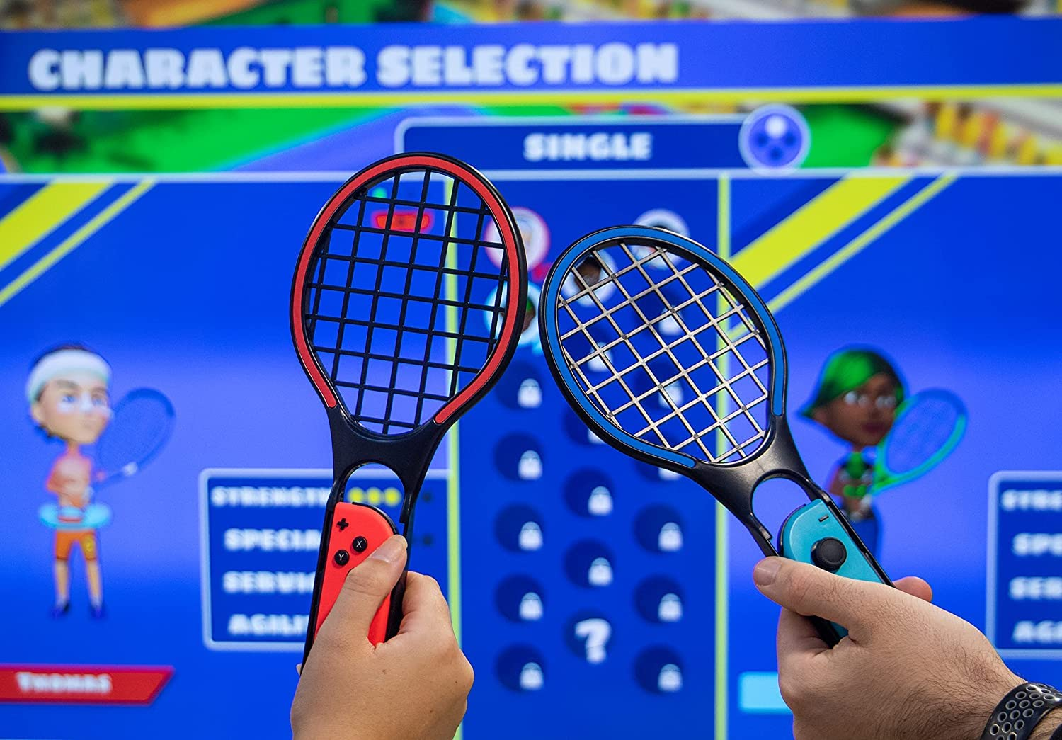 Manos sosteniendo mandos rojos y azules con forma de raqueta de tenis frente a una pantalla que muestra la selección de personajes de un juego de tenis.
