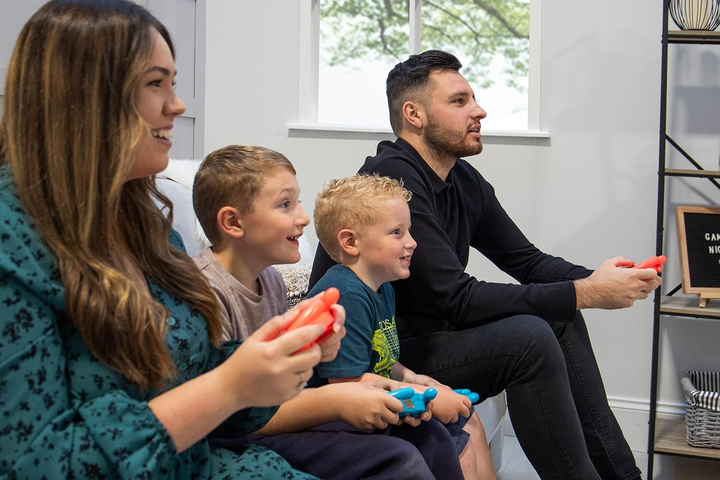 Una familia de cuatro está jugando un videojuego. Sostienen mandos y sonríen. Un letrero dice 'Noche de juegos'.