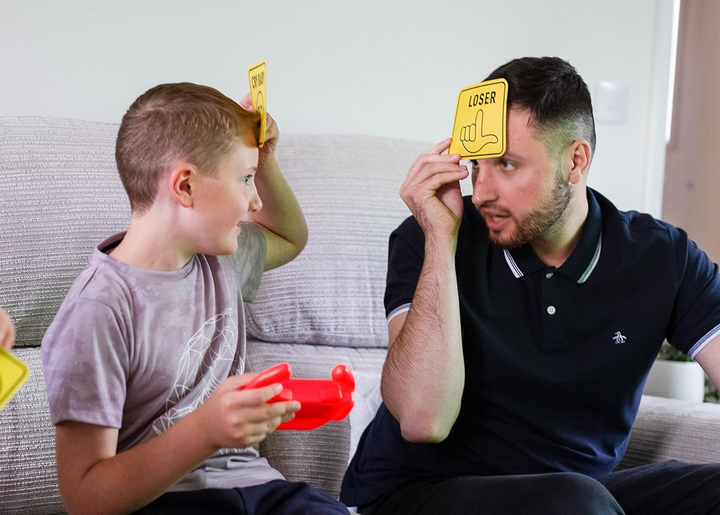 Un niño y un hombre juegan. Ambos sostienen cartas sobre sus cabezas. La carta del hombre dice 'Perdedor', la del niño 'Llorón'.