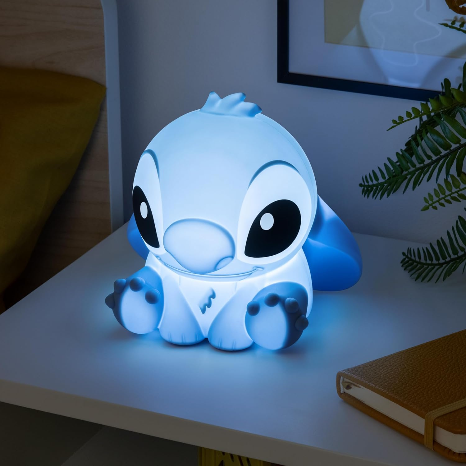 Egy kék Stitch éjszakai fény egy fehér felületen ül. Hátul egy jegyzetfüzet, egy növény és egy bekeretezett kép látható.
