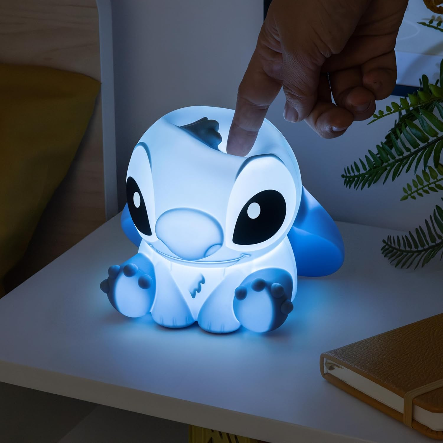 Egy ujj megérinti a kék színben világító Stitch lámpát. A lámpa egy fehér asztalon van egy jegyzetfüzet és egy növény mellett.
