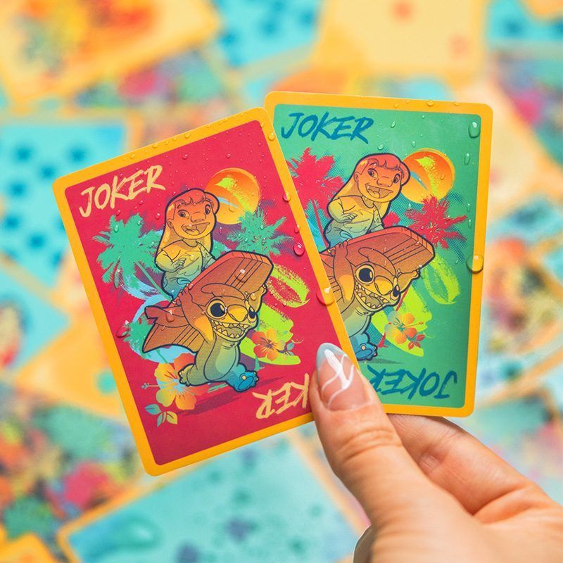 Kézben két Joker kártya, rajzfilmfigurákkal. A piros kártyán Stitch karakter, a zöldön egy másik.