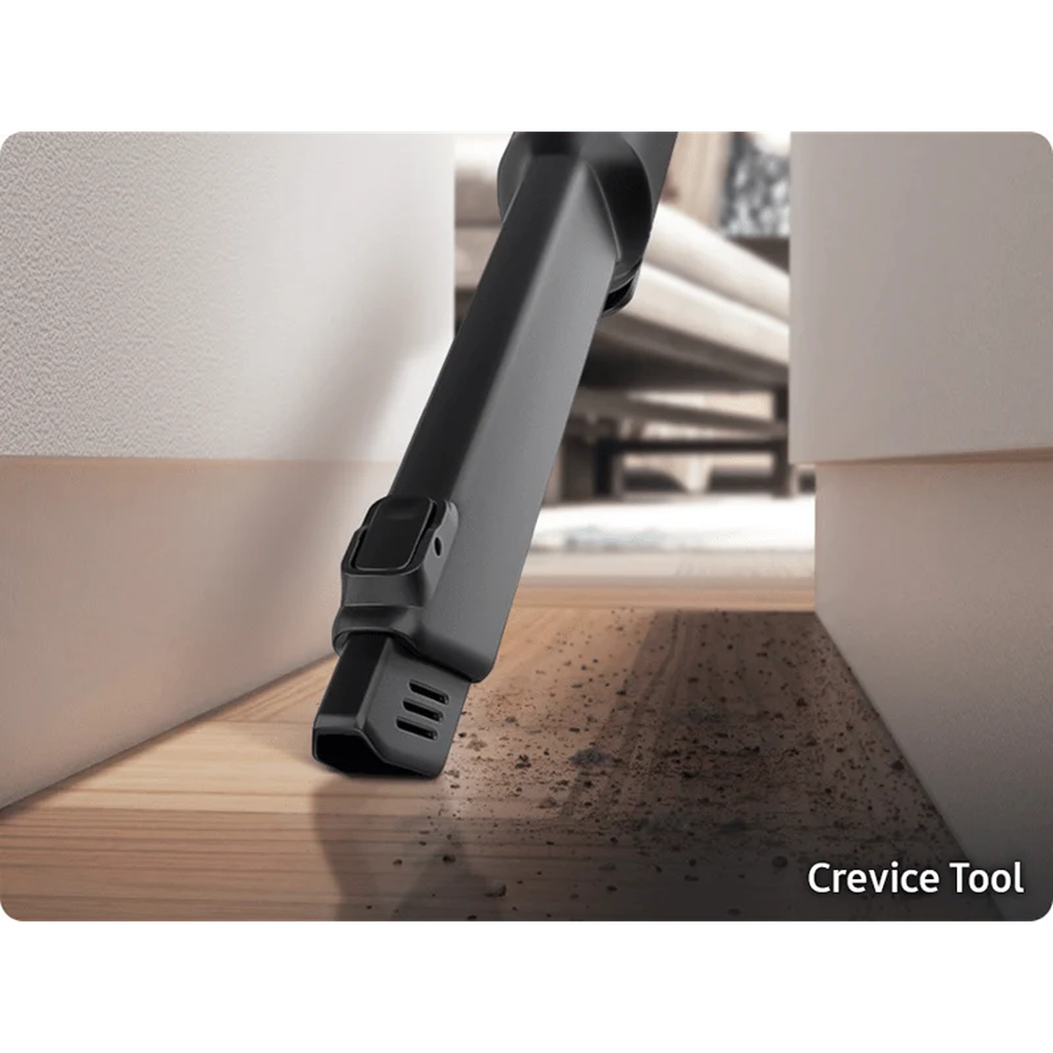 A black crevice tool cleaning dust on a wooden floor. The tool is between two white walls. The background is blurred.