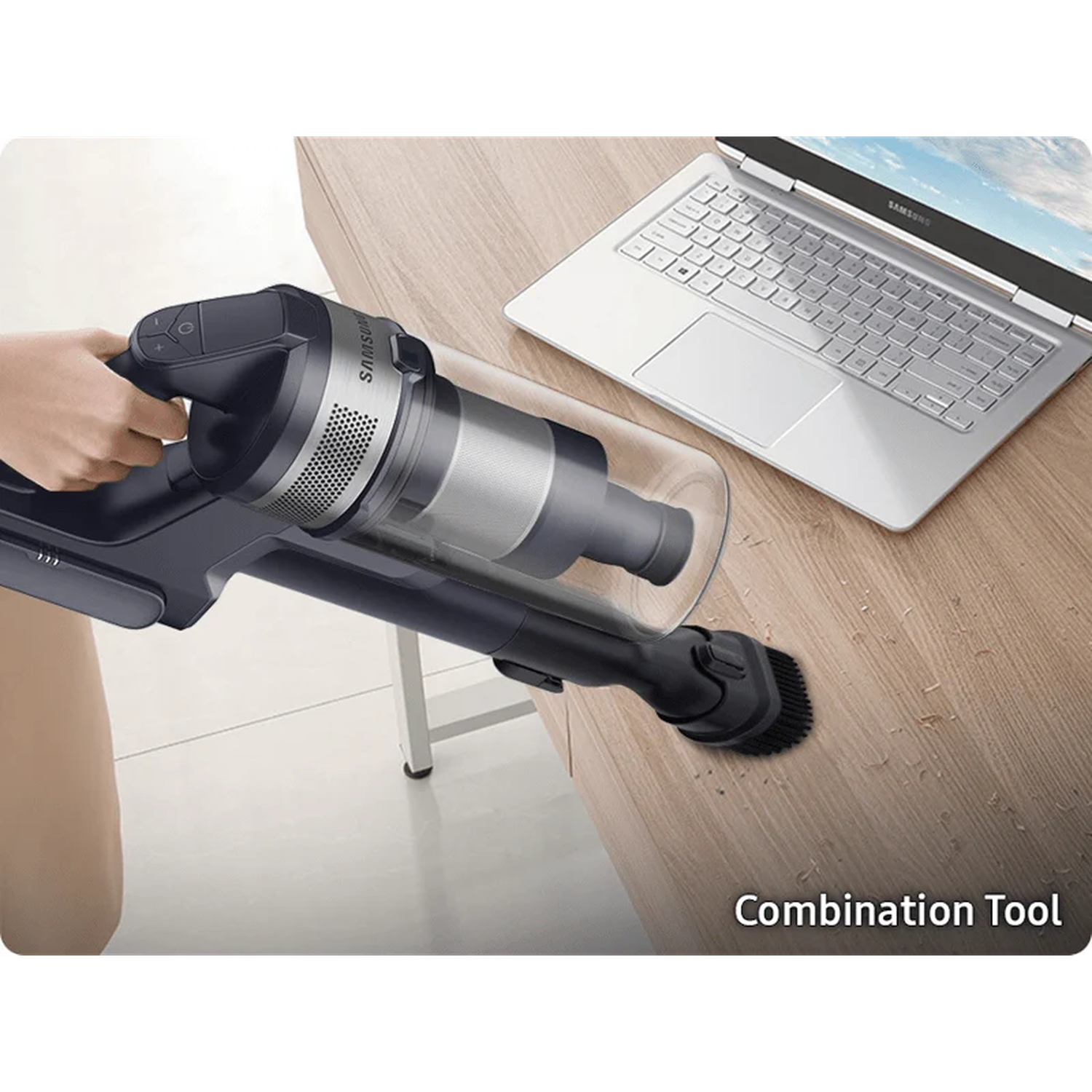 A person uses a vacuum cleaner on a wooden table with a laptop. The vacuum is black and silver, and the laptop is silver.