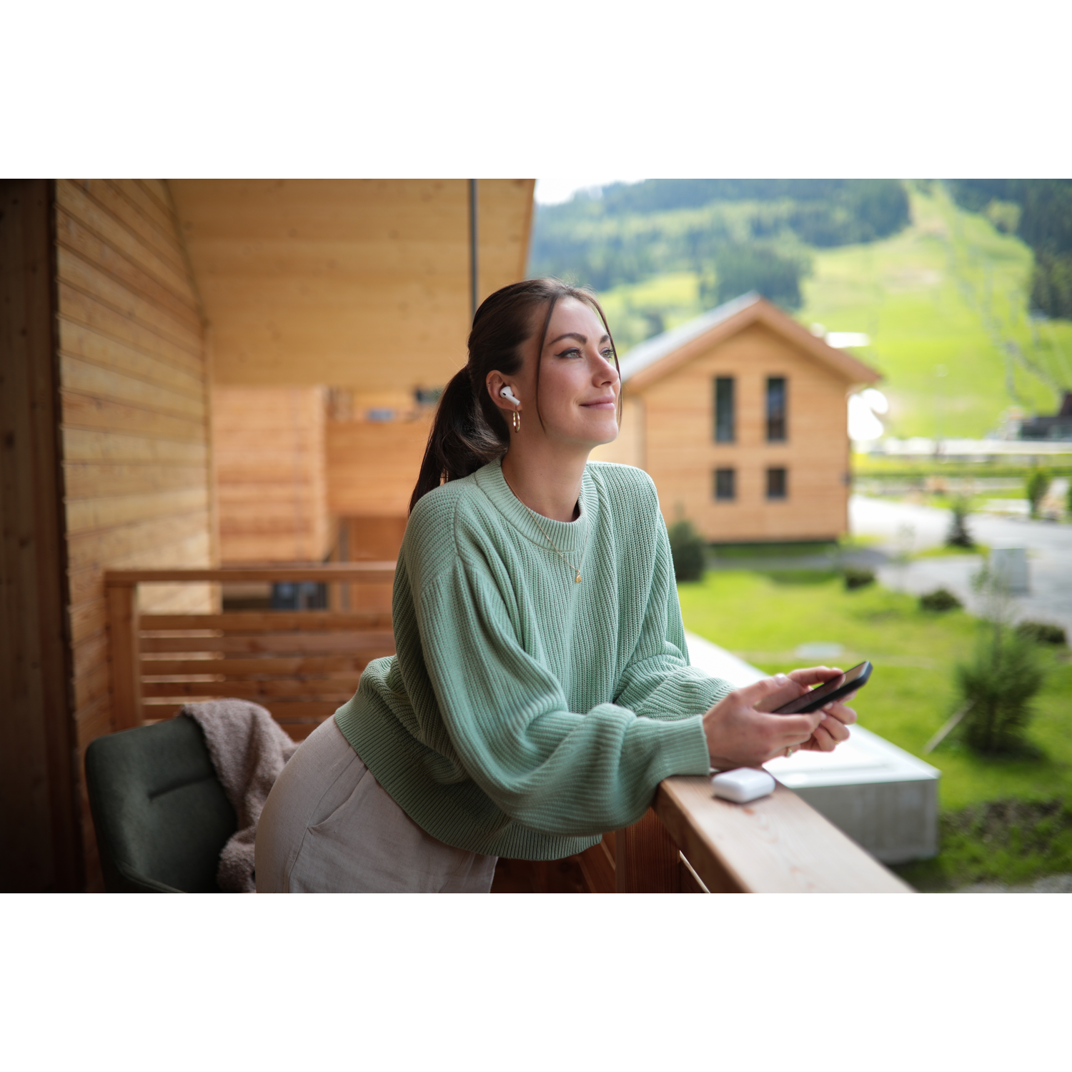 Woman on a balcony wearing earbuds, holding a phone, looking up. Background shows wooden houses and a mountain.
