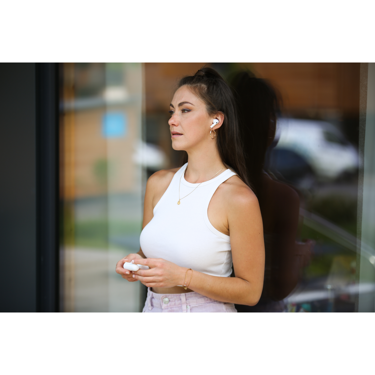 A woman with long dark hair, wearing a white top and pink pants, holds white earbuds, stands by a glass wall.