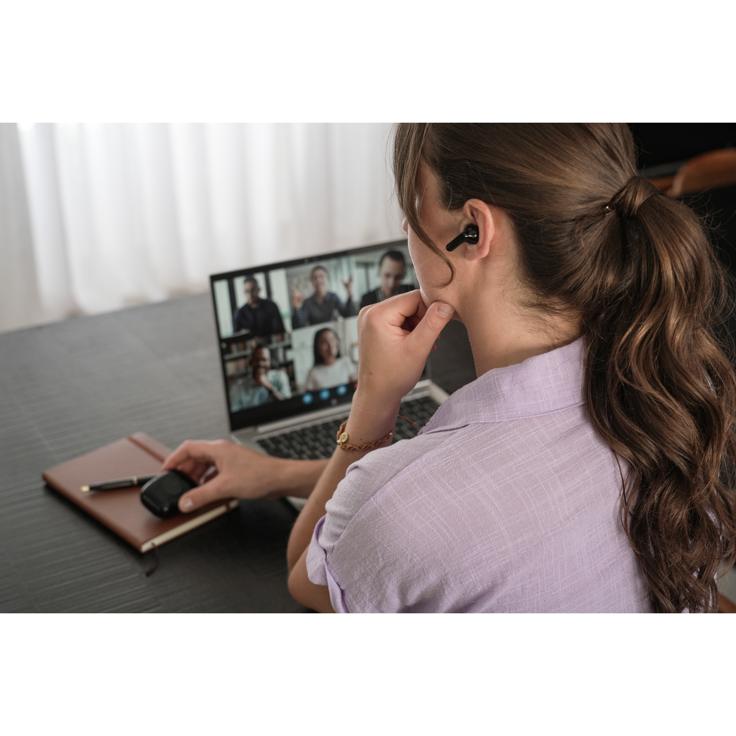 Een vrouw met oordopjes neemt deel aan een videogesprek op een laptop. Ze houdt een muis vast en raakt haar gezicht aan. Een notitieboek en een pen liggen op tafel.
