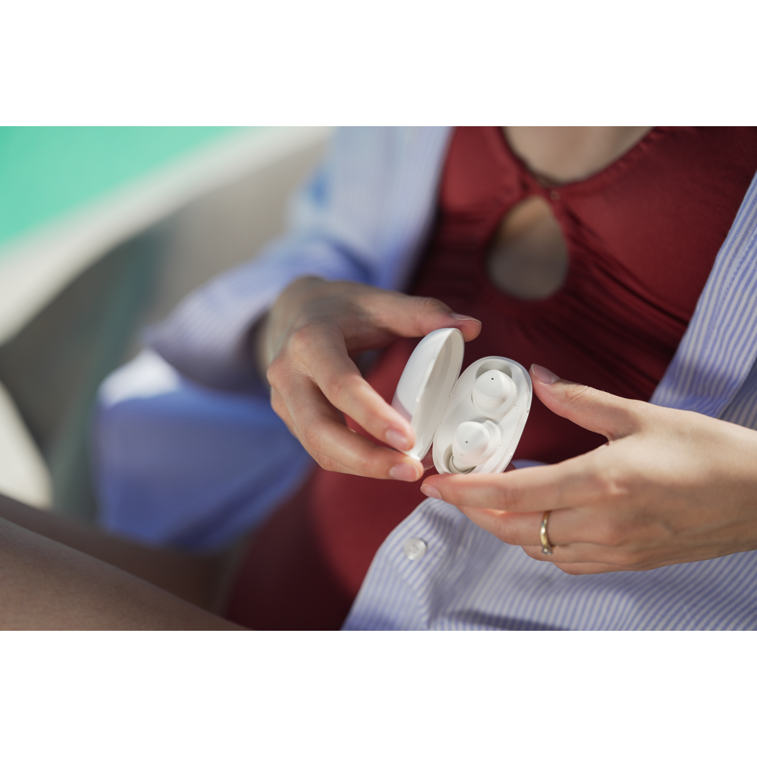 Une personne tient un étui d'écouteurs blanc avec des écouteurs. Elle porte un maillot de bain rouge et une chemise à rayures bleues. Une bague en or est visible.
