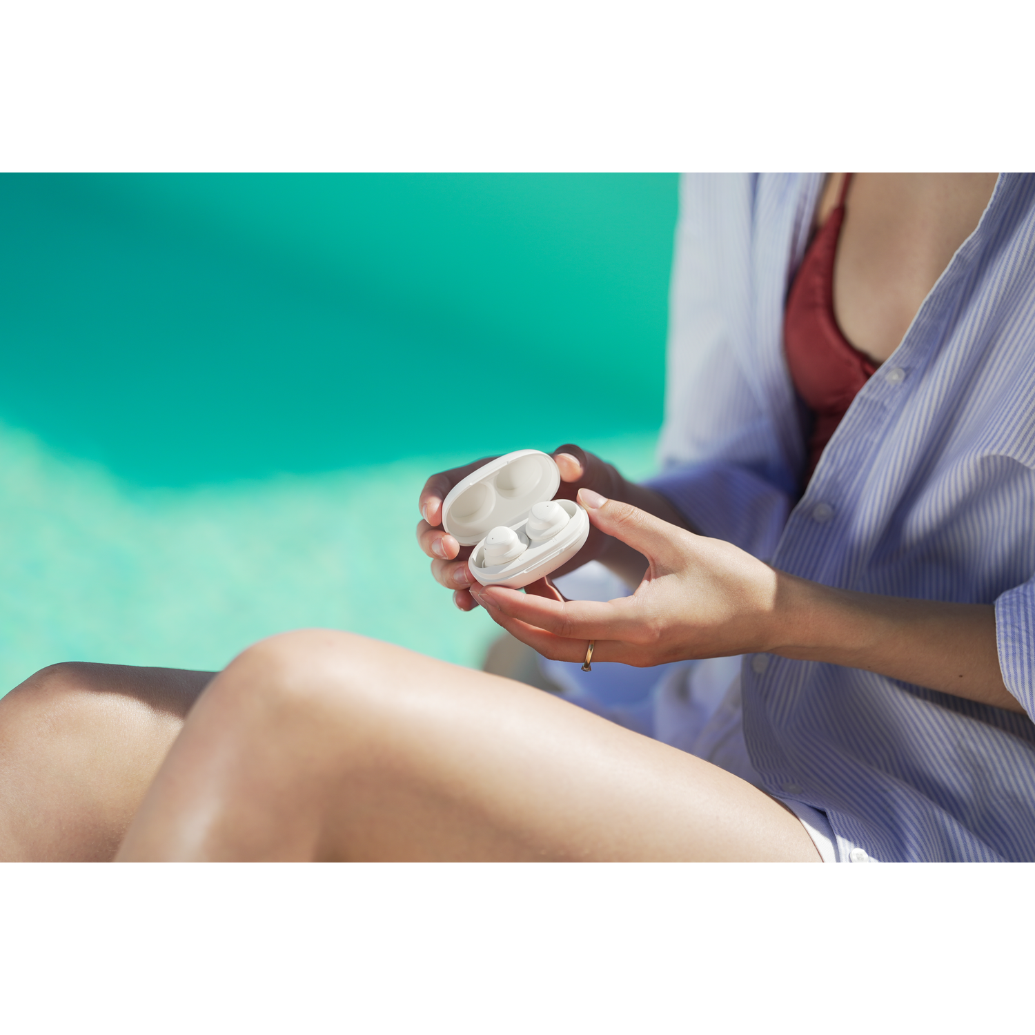 Une personne tenant un étui d'écouteurs blanc ouvert. Elle porte une chemise rayée bleue et un haut bordeaux, avec une piscine turquoise en arrière-plan.