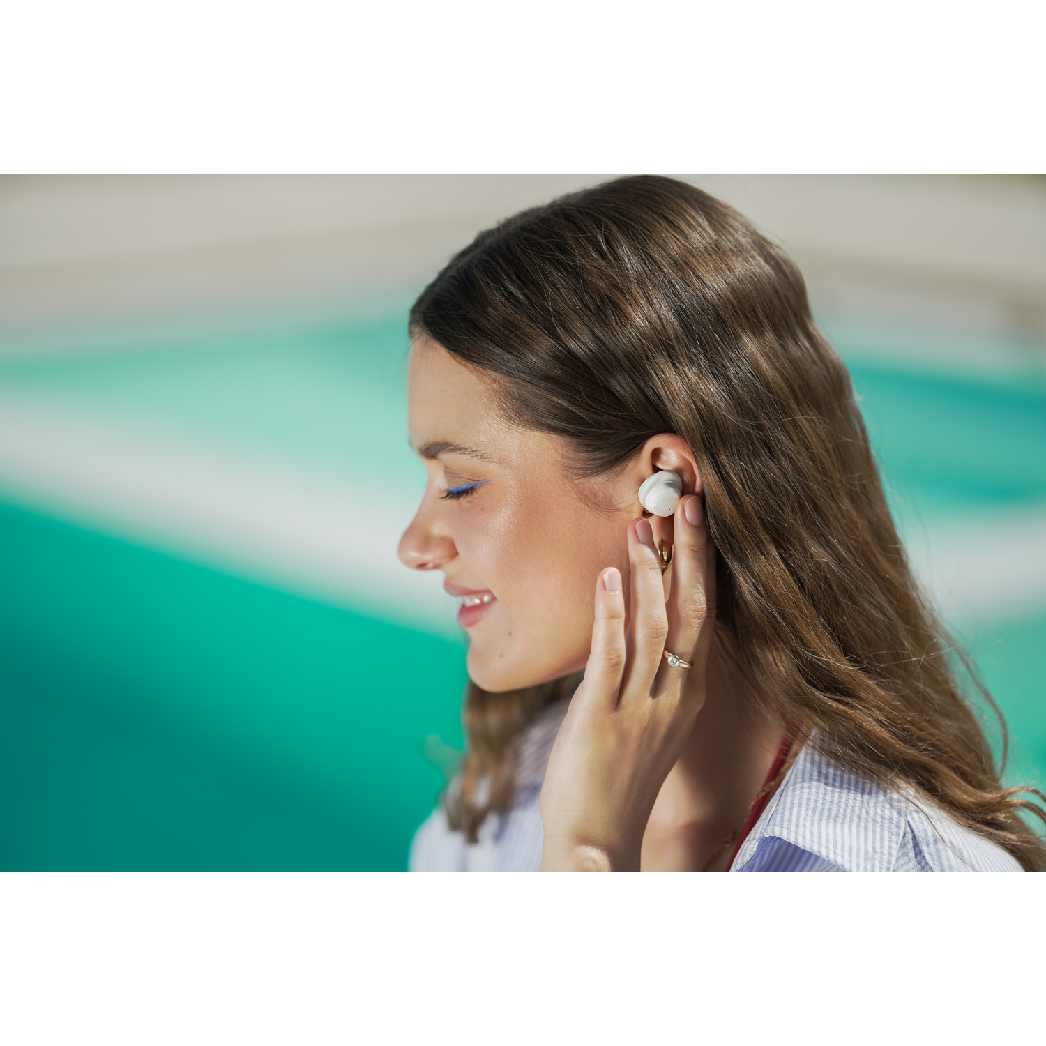 Une femme aux longs cheveux bruns portant un écouteur. Elle a un eye-liner bleu, une bague et porte une chemise rayée bleue et blanche.