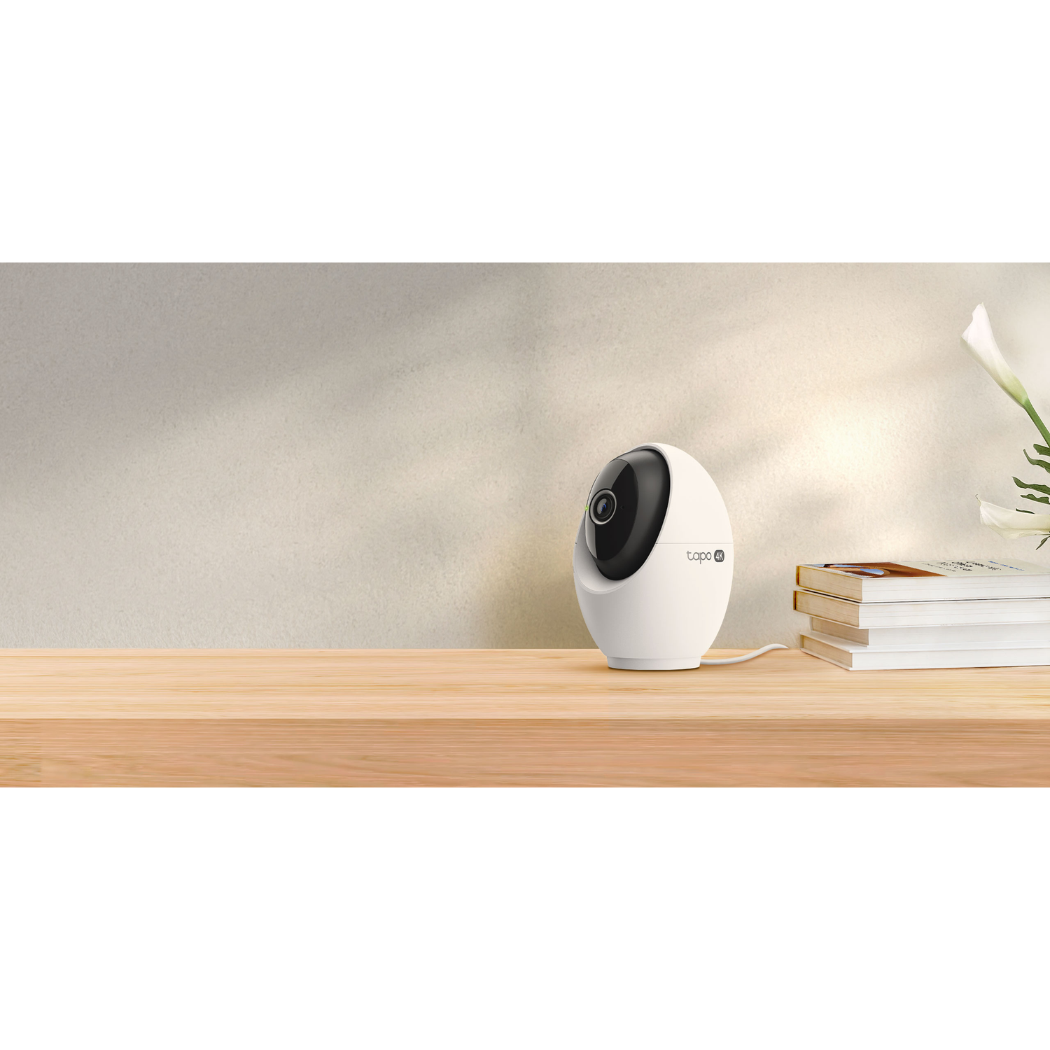 A white Tapo security camera sits on a wooden table. Beside it, a stack of books and a white flower. Beige wall in the background.
