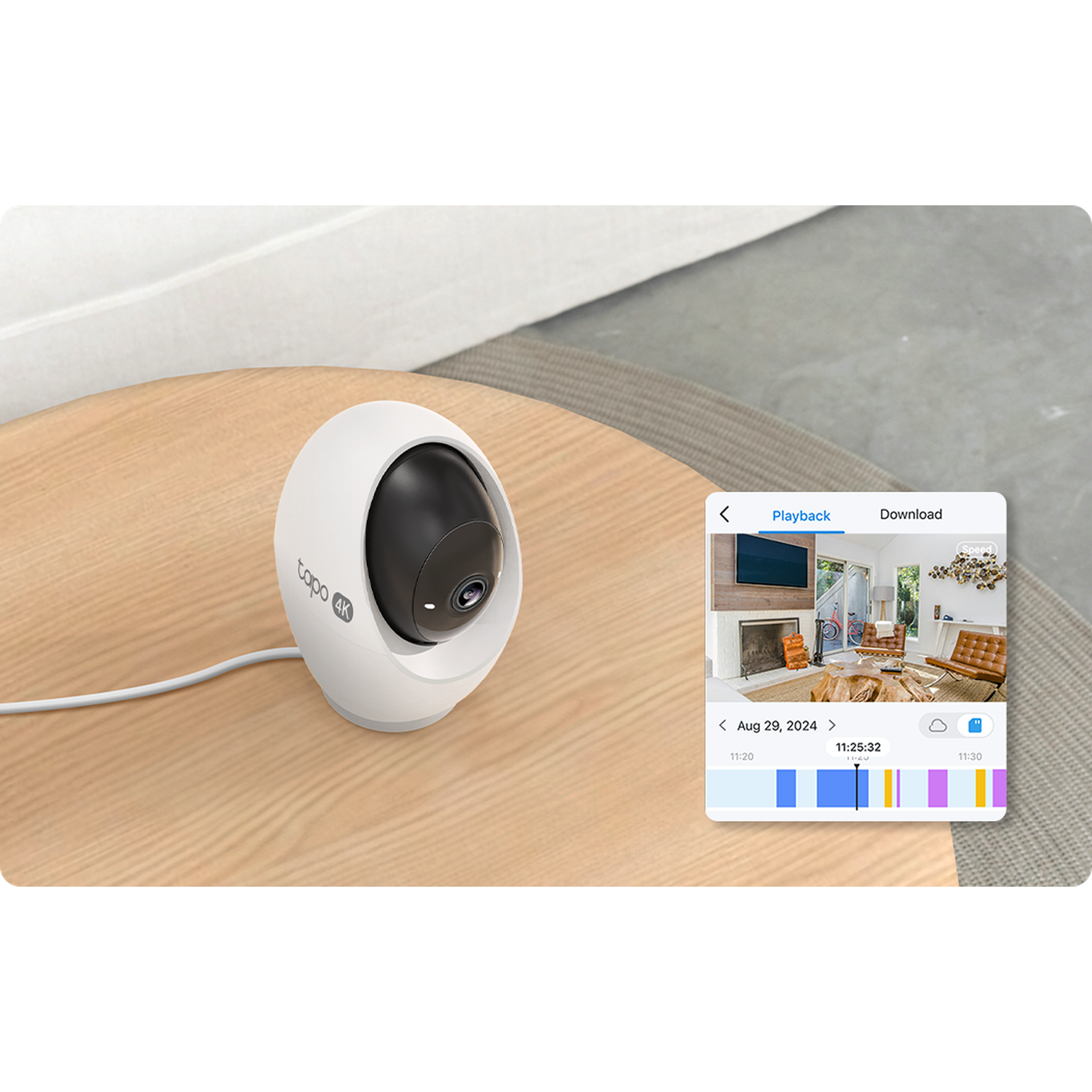 A white and black security camera sits on a wooden table. A screen shows a room and a timeline of video playback.