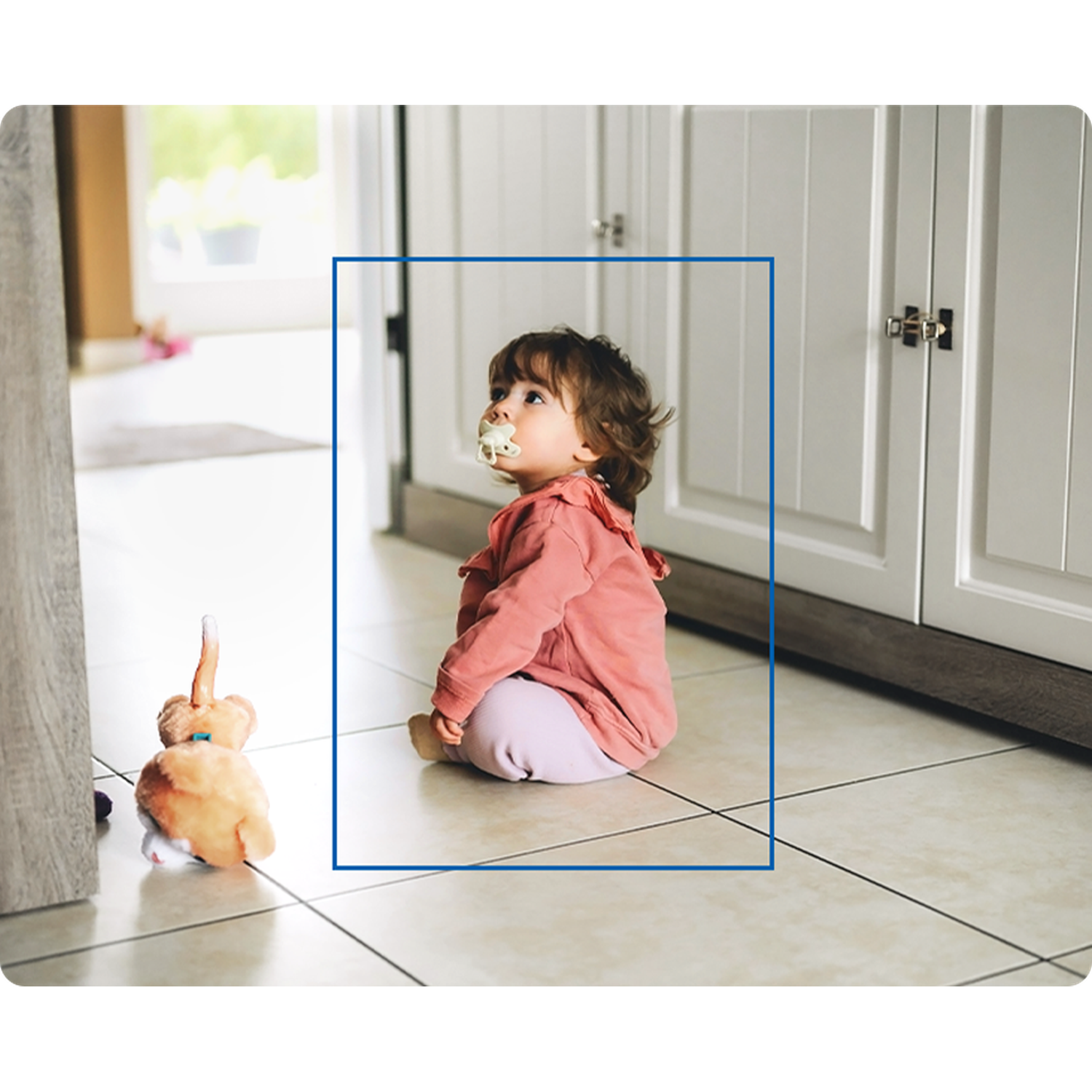 A baby sits on a tiled floor, wearing a pink shirt and light pants, looking up with a pacifier in its mouth. There's a toy nearby.