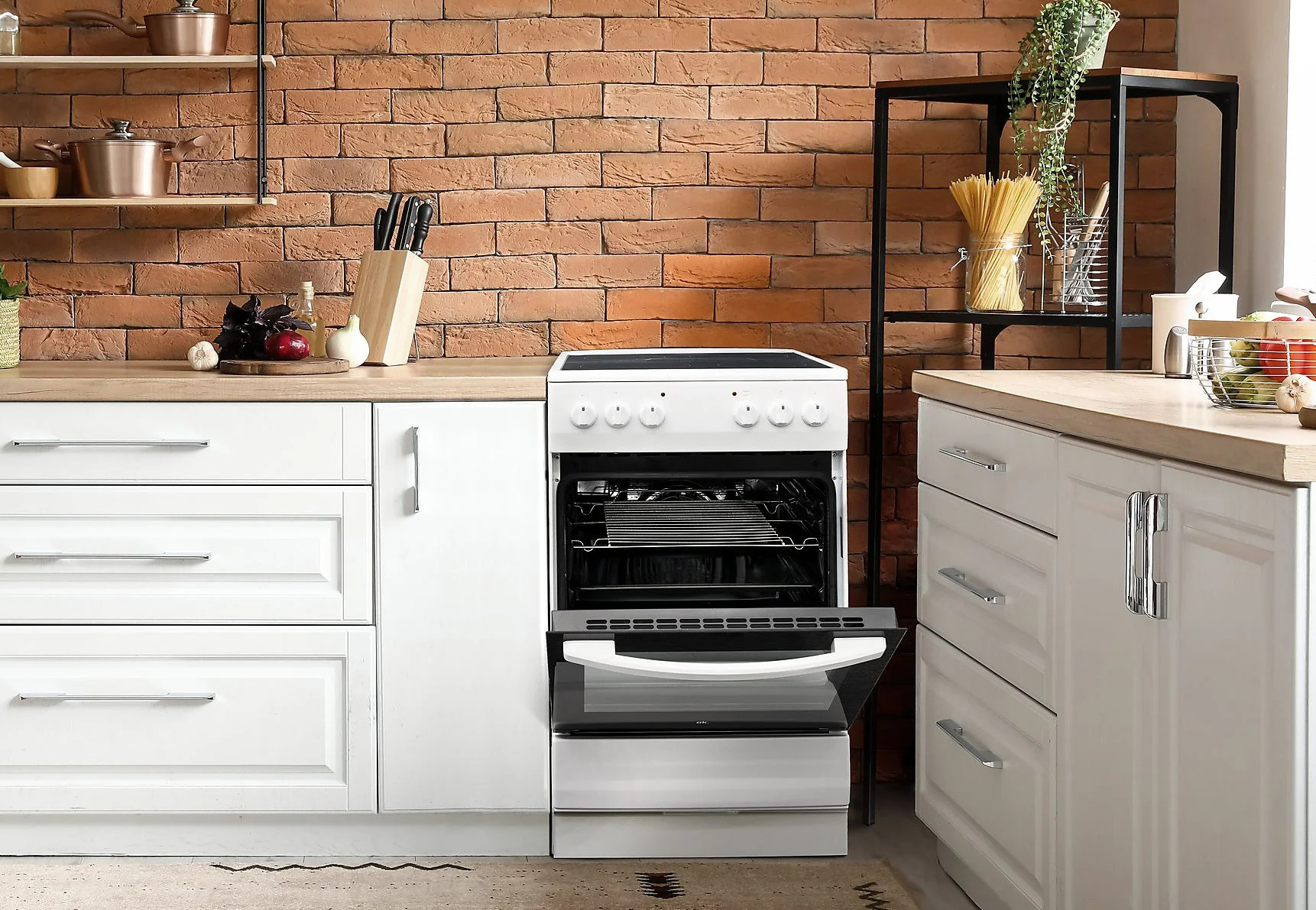 Kuchnia z białym piekarnikiem, ceglaną ścianą i szafkami. Drzwi piekarnika są otwarte. Na blacie owoce i noże. Makaron na półce.