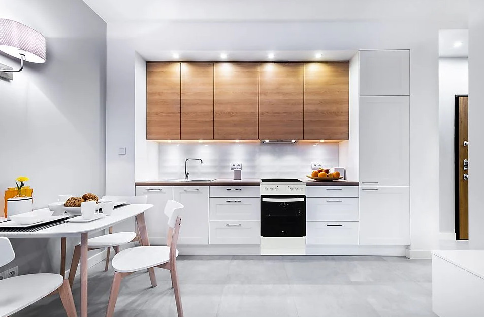 Nowoczesna kuchnia z białymi szafkami, drewnianymi szafkami górnymi i stołem jadalnym z krzesłami. Widoczny piekarnik i zlew.