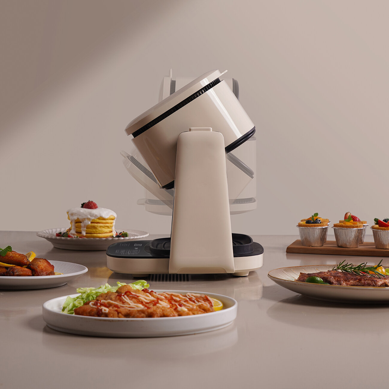 Beżowy robot kuchenny stoi obok stołu z różnymi potrawami: naleśnikami, smażonym kurczakiem, stekiem i babeczkami.