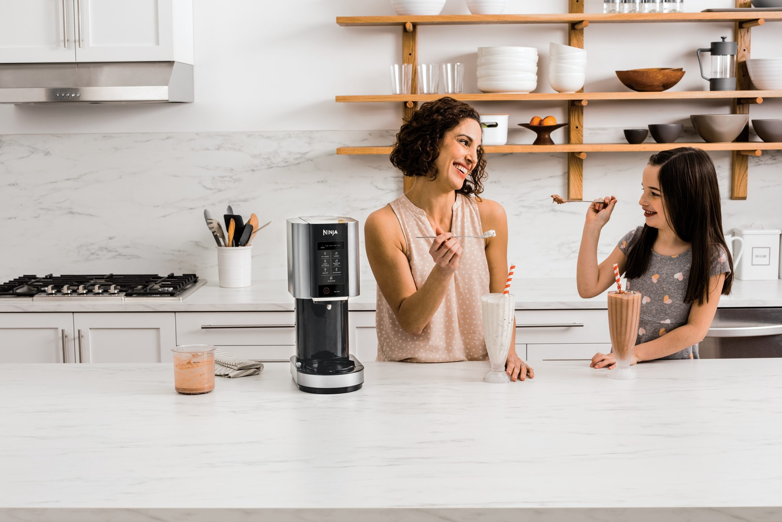 Kobieta i dziewczynka w kuchni dzielą się koktajlem mlecznym. Na blacie stoi blender Ninja. Półki zawierają miski i pojemnik na cukier.