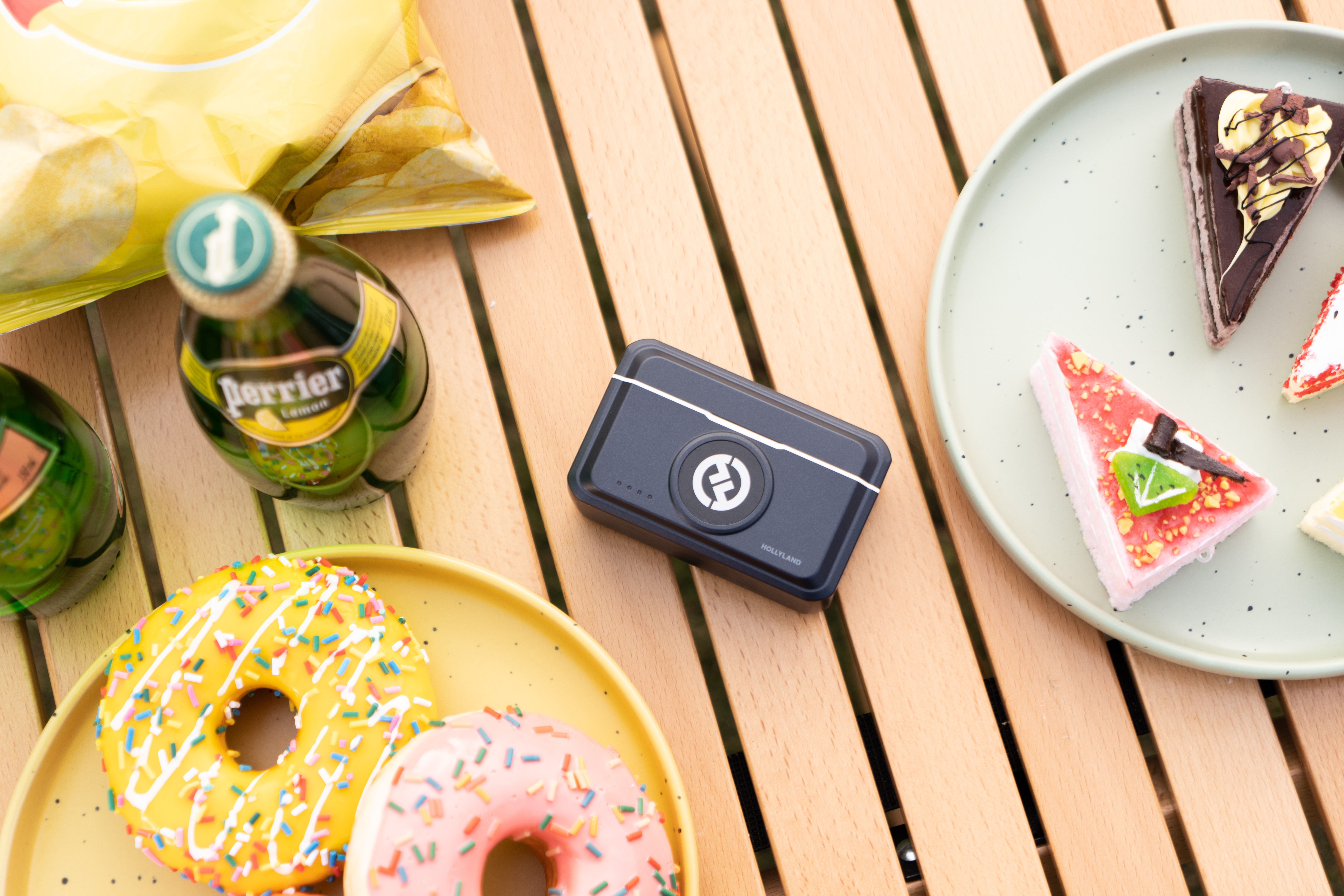 Stół z pączkami, ciastem, butelkami Perrier, chipsami i czarnym urządzeniem. Pączki są żółte i różowe.