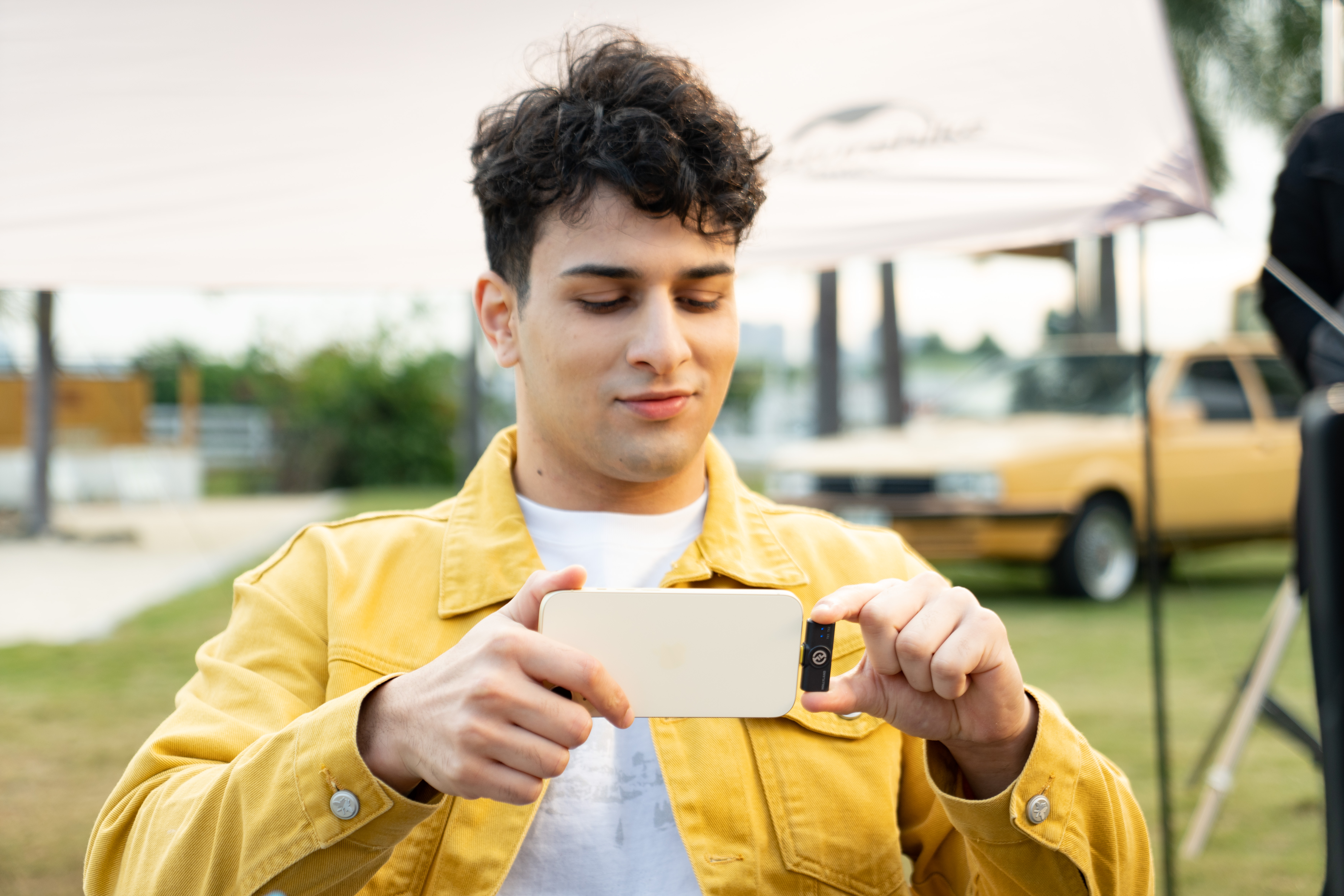 Mężczyzna w żółtej kurtce trzyma telefon, patrząc na niego. Żółty samochód i biały namiot w tle.