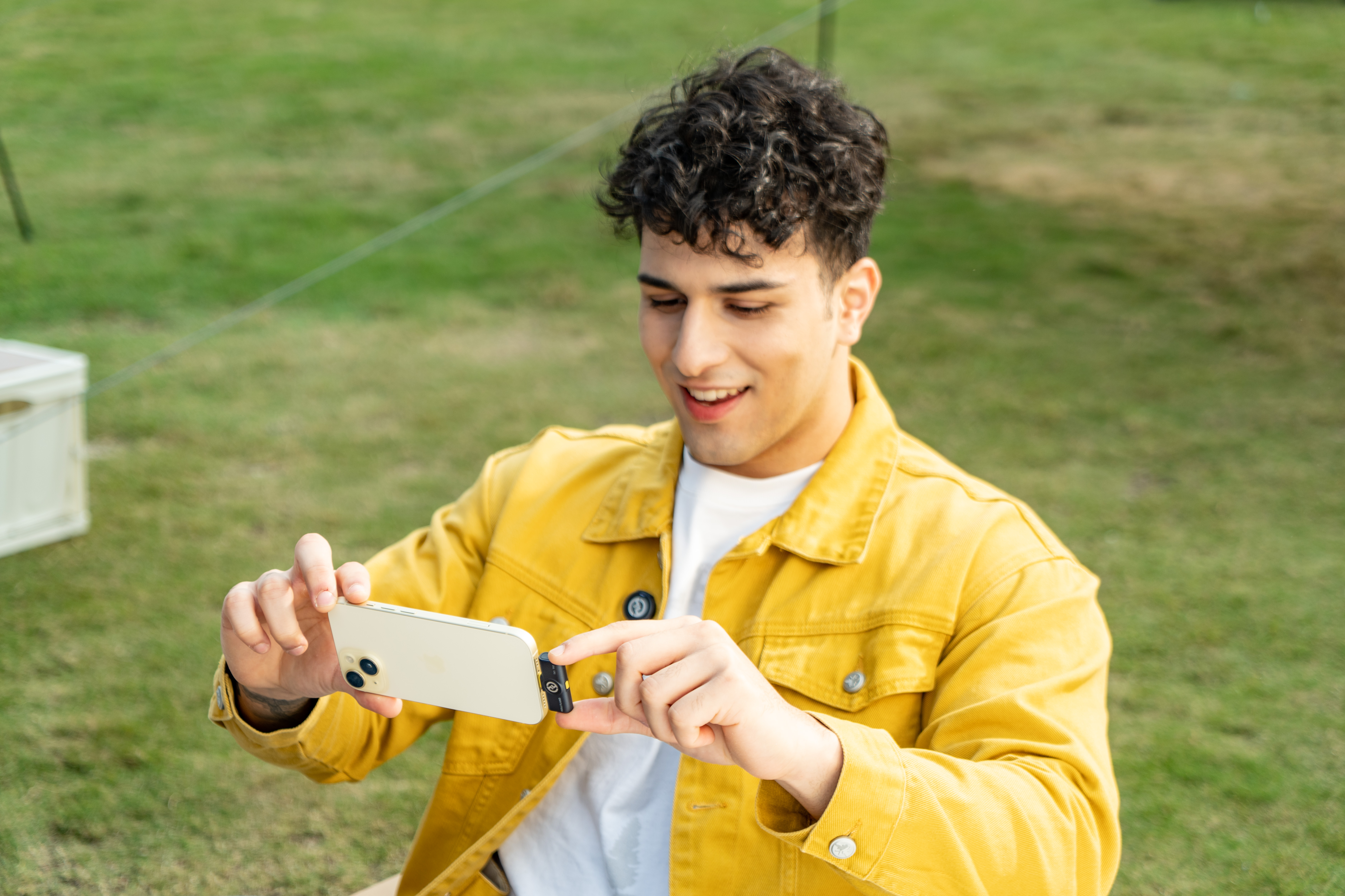 Mężczyzna w żółtej kurtce trzyma telefon, uśmiechając się. Zielona trawa w tle. Mocuje mały mikrofon.