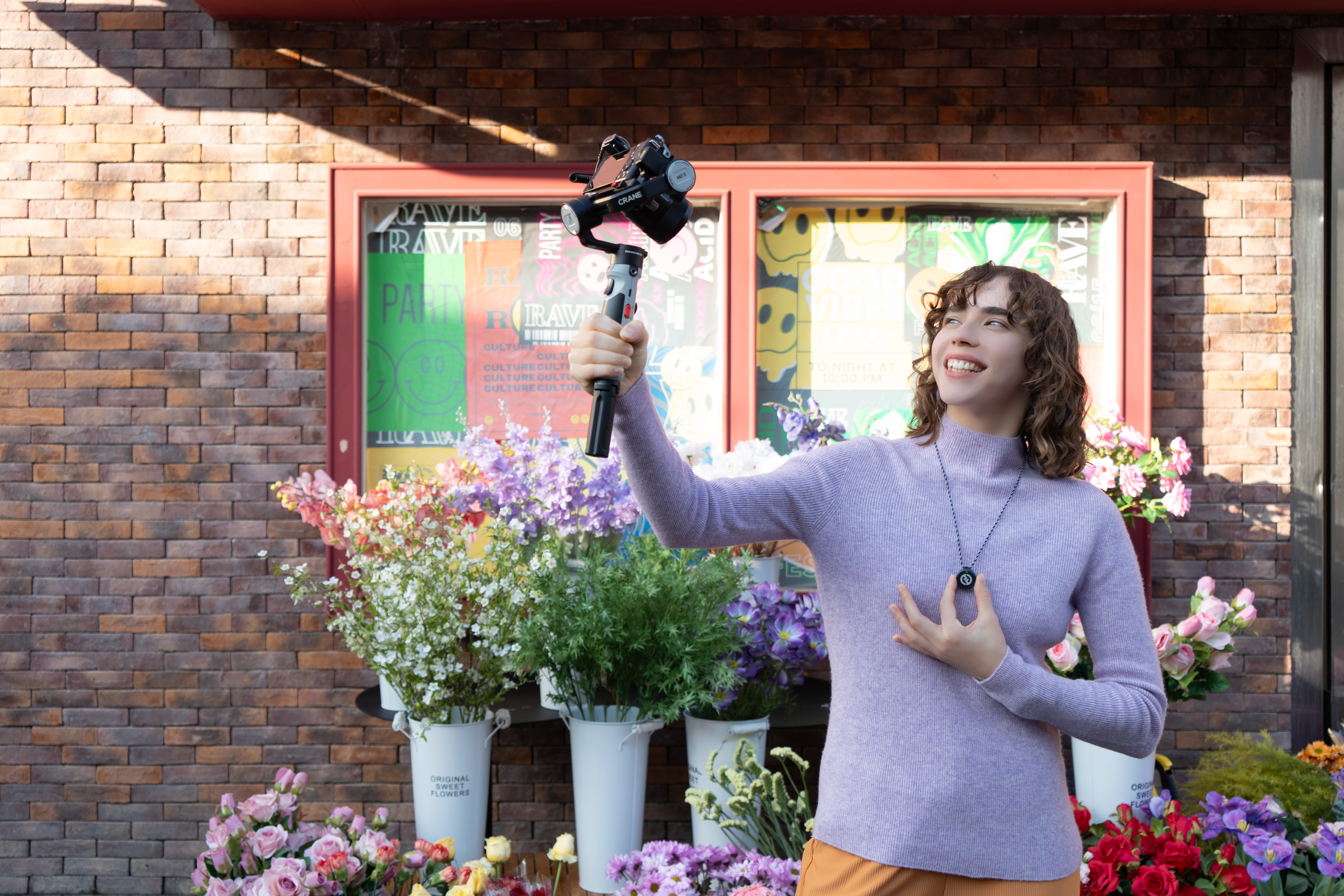Młoda kobieta z aparatem, uśmiechnięta, otoczona kwiatami. Tło: ceglana ściana, plakaty i naszyjnik.