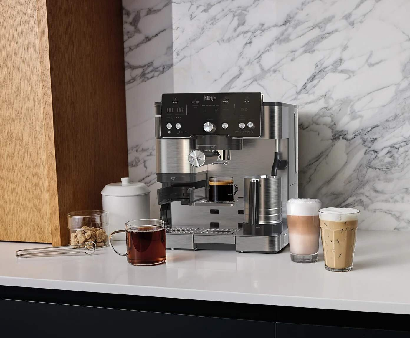 Ekspres do kawy Ninja z filiżankami kawy i latte na białej powierzchni. Ponadto szklanka herbaty i brązowy cukier.