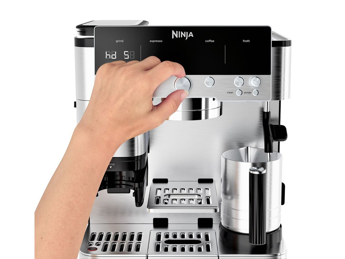 Ręka obraca pokrętło na srebrnym ekspresie do kawy Ninja. Wyświetlacz pokazuje liczby. Widoczny jest dzbanek do spieniania mleka.
