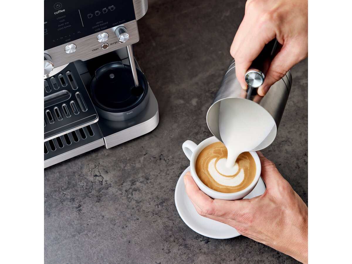 Osoba wlewa mleko do filiżanki kawy, tworząc latte art. Po lewej stronie ekspres do kawy. Powierzchnia jest ciemnoszara.