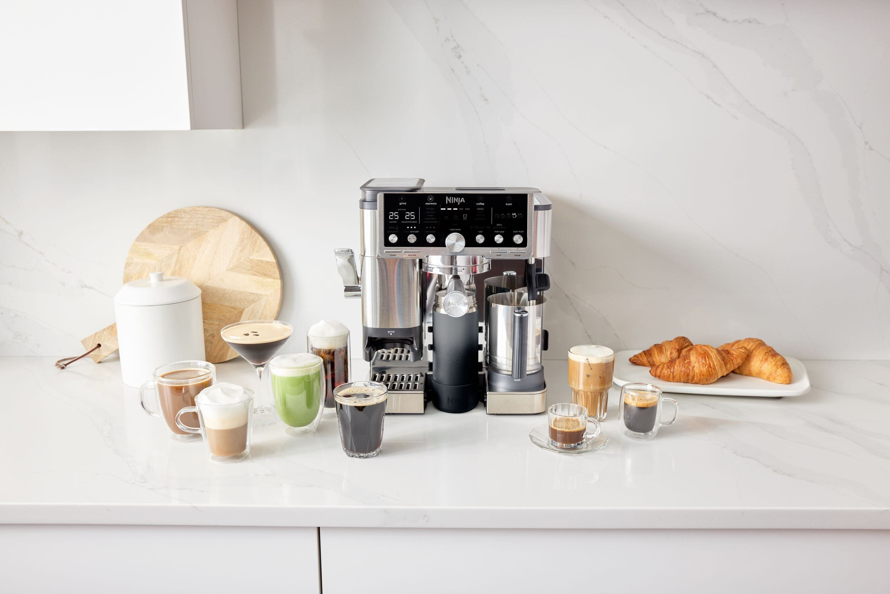 Ekspres do kawy z różnymi napojami kawowymi i rogalikami na białym blacie. Napoje obejmują espresso, latte i kawę mrożoną.