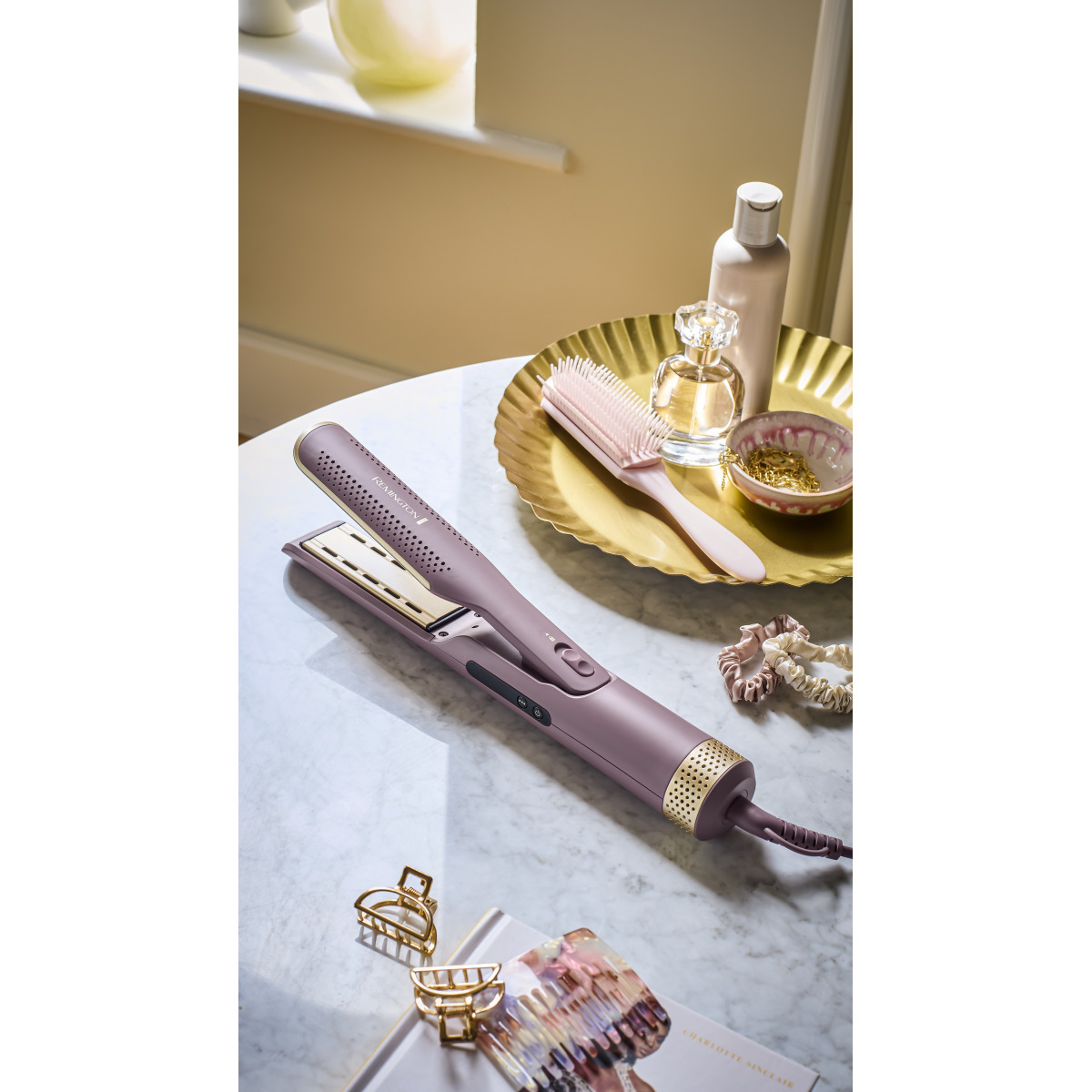 Prostownica, szczotka, perfumy, gumki do włosów i spinki do włosów leżą na marmurowym stole. Prostownica jest fioletowo-złota.