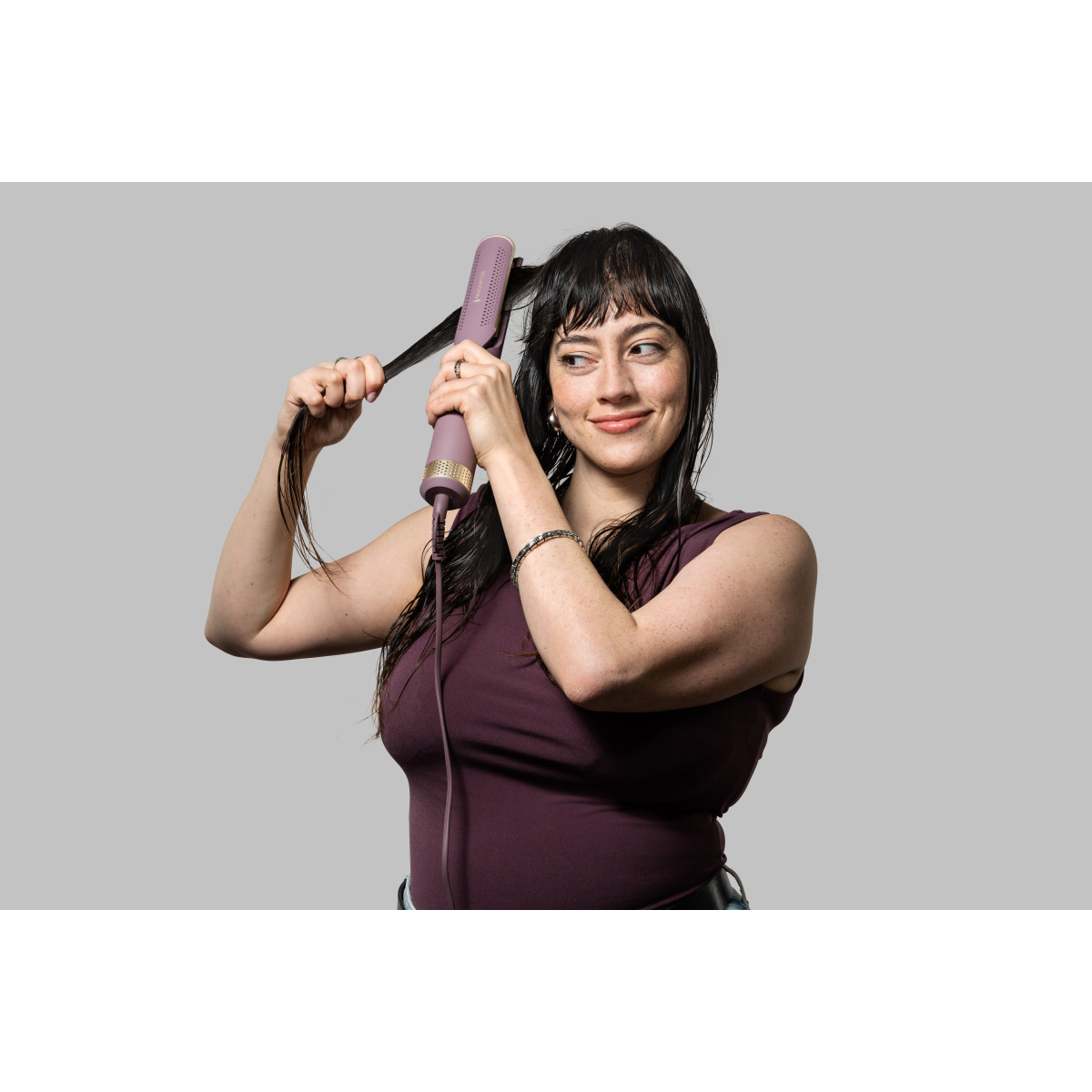 Kobieta używa fioletowo-złotej prostownicy do ciemnych włosów. Ma na sobie bordowy top i patrzy w bok z uśmiechem.