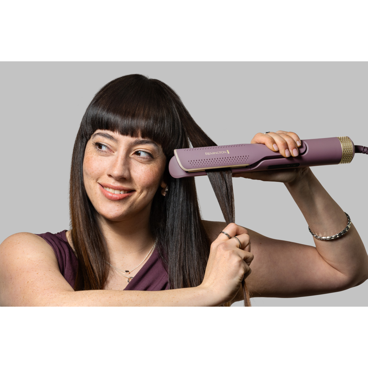 Kobieta używa fioletowej prostownicy do włosów. Ma ciemne włosy i uśmiecha się. Tło jest szare.