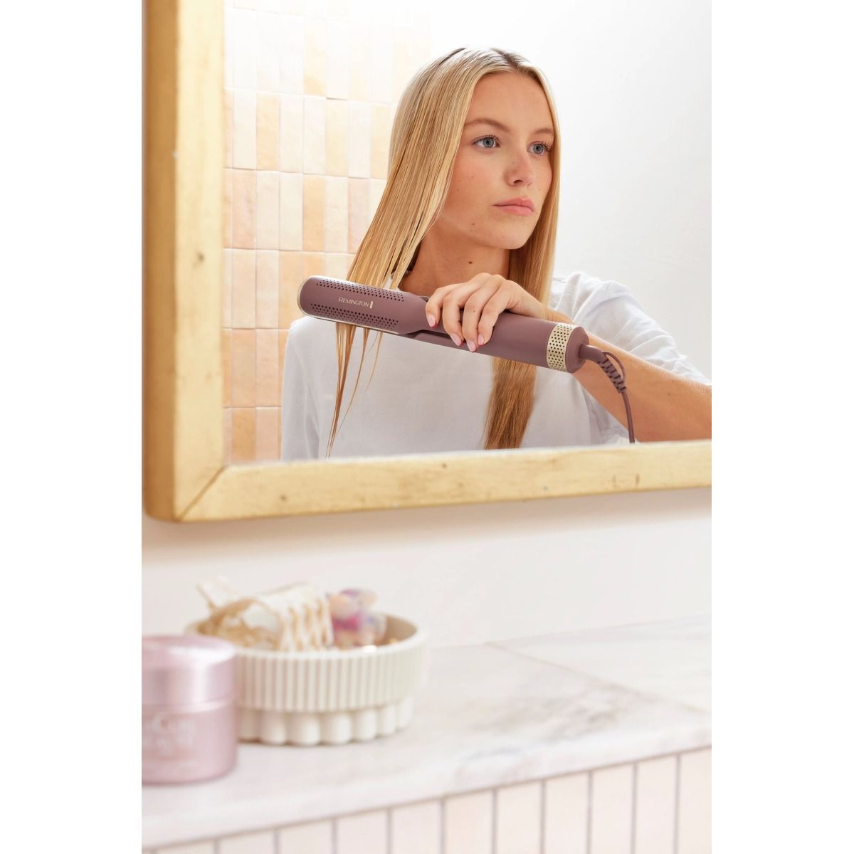 Kobieta z blond włosami używa prostownicy przed lustrem. Widoczny pojemnik na kosmetyki i akcesoria.