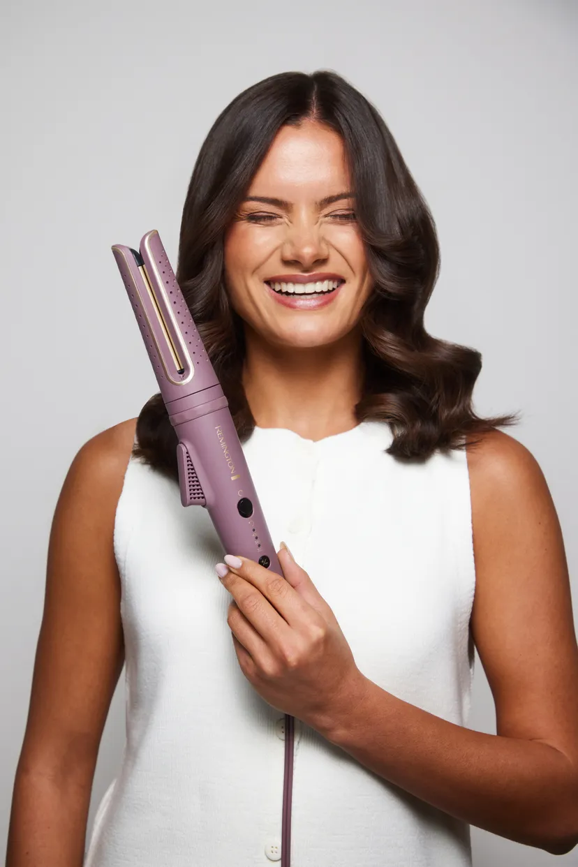 Kobieta uśmiecha się, trzymając fioletową lokówkę Remington. Ma na sobie biały top, ciemne włosy i jasne tło.