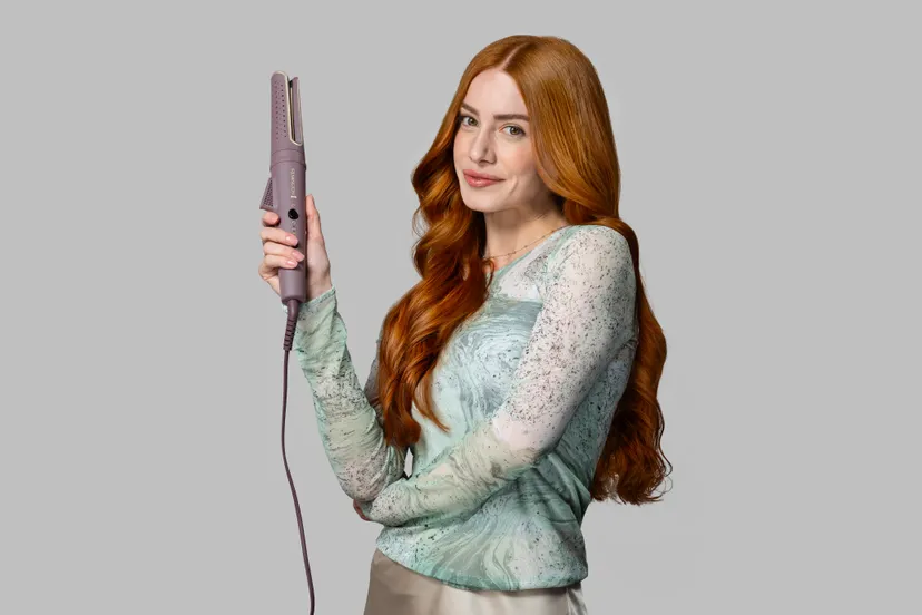 Kobieta z rudymi włosami trzyma prostownicę. Ma na sobie zieloną koszulę. Tło jest szare.