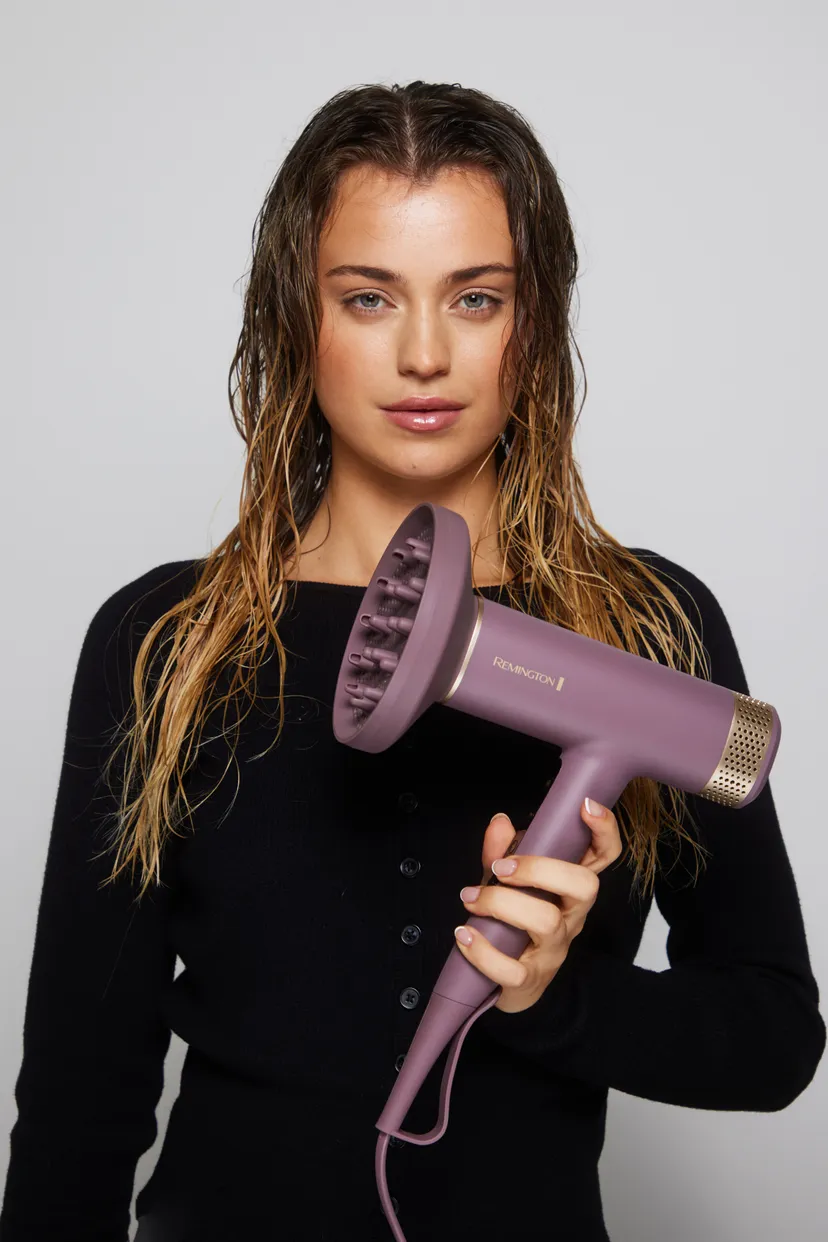 Kobieta z mokrymi włosami trzyma fioletową suszarkę do włosów Remington. Ma na sobie czarny sweter i jasną skórę. Tło jest białe.
