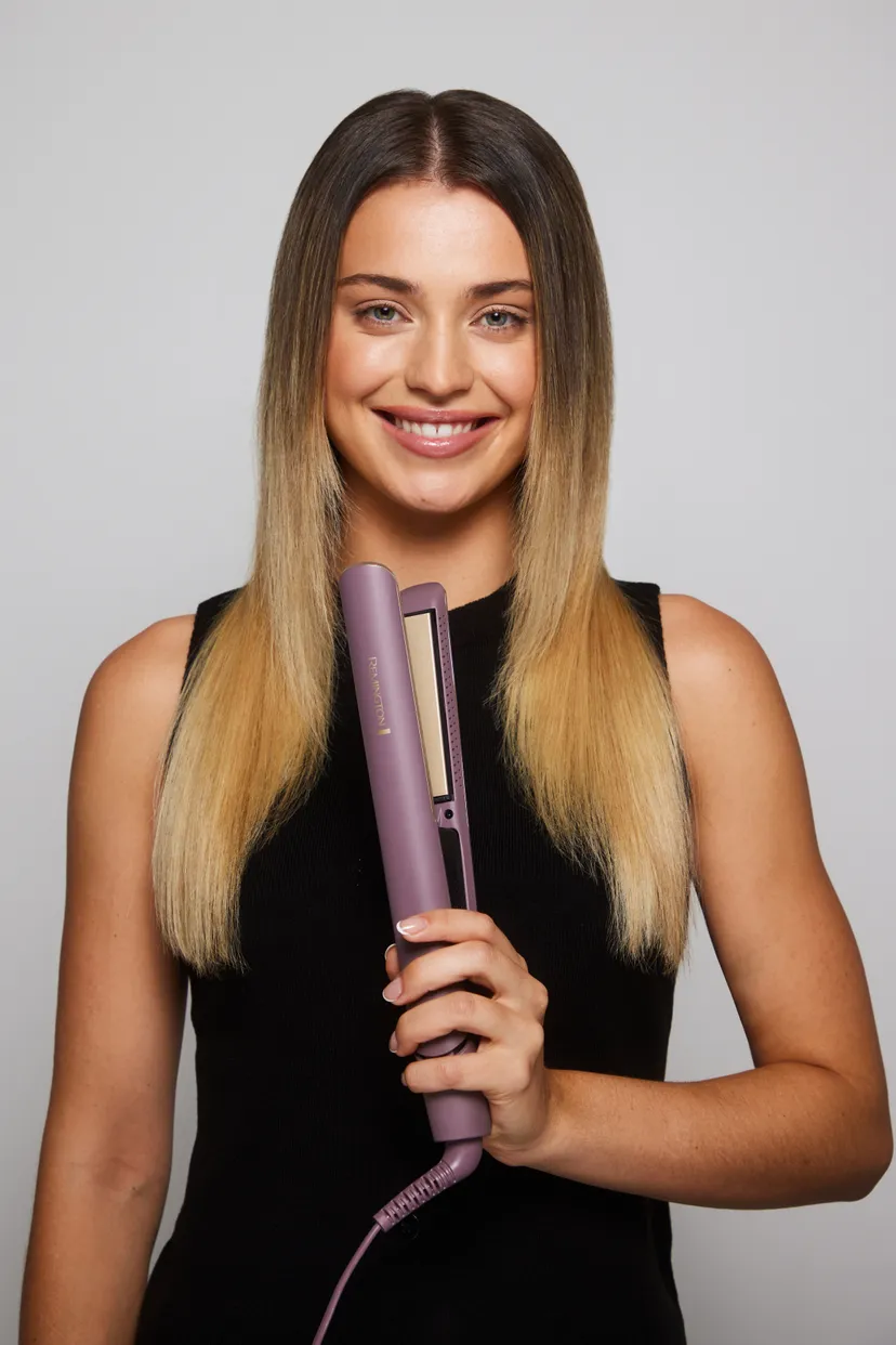 Kobieta z blond włosami trzyma fioletową prostownicę do włosów. Uśmiecha się do kamery, mając na sobie czarny top.